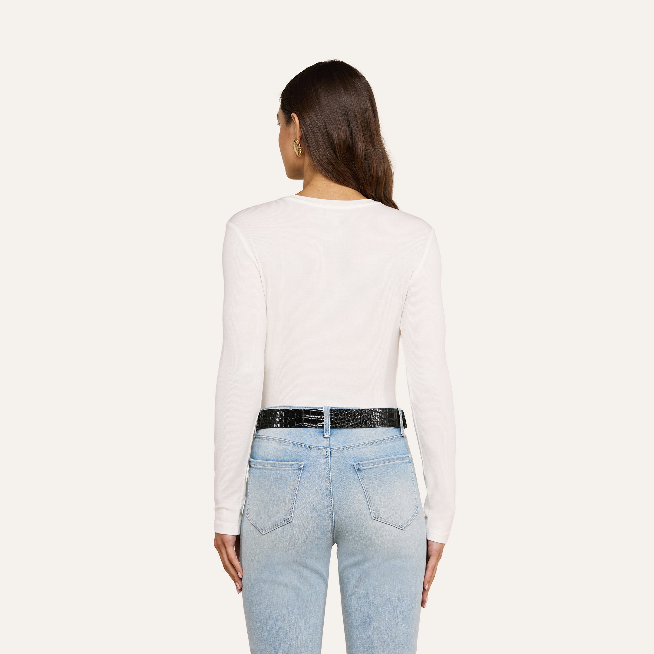 A woman with long brown hair is shown from the back, wearing the Tess Long Sleeve Tee in white, paired with light blue jeans and a black belt, standing against a plain light background.