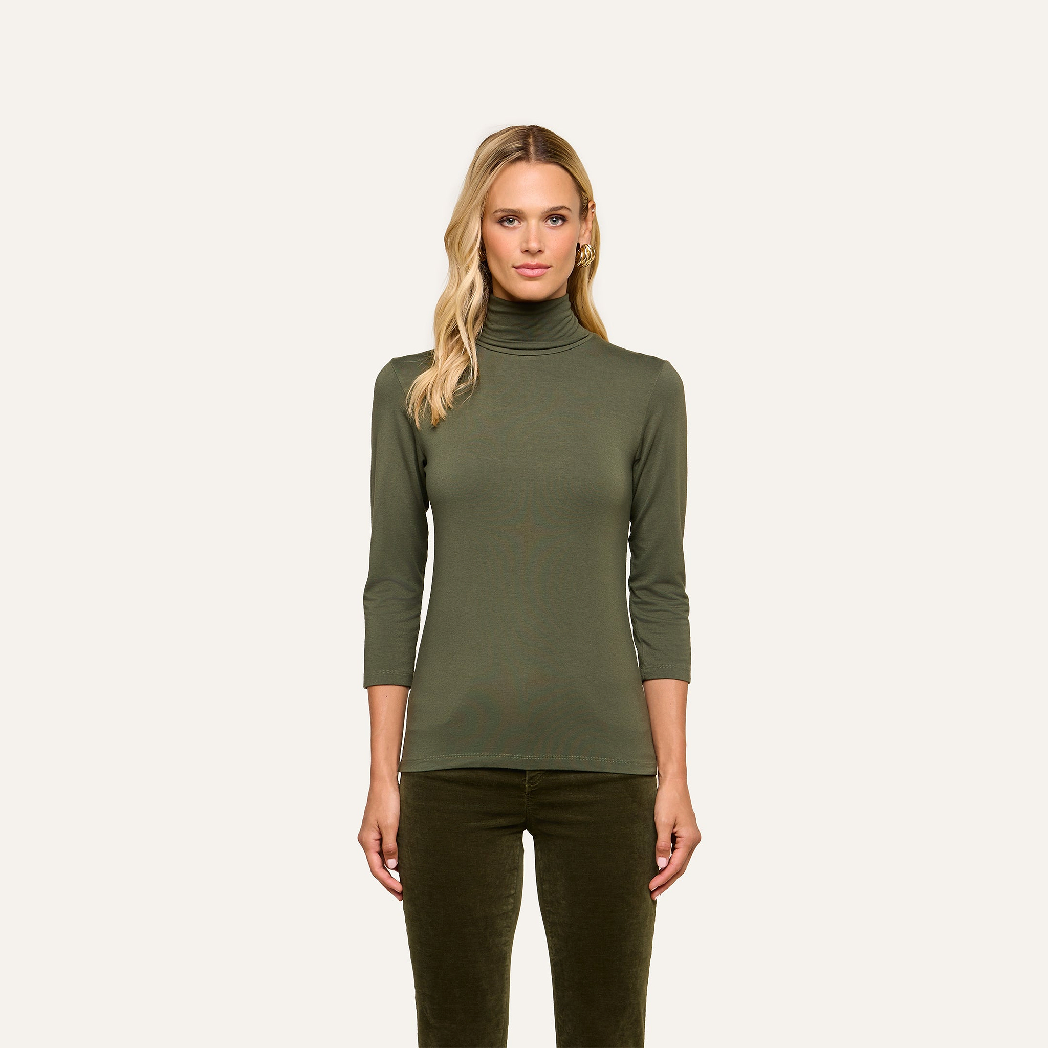A woman wearing the Aja Turtleneck—a slim fit olive green top with three-quarter sleeves—along with matching pants stands facing forward against a plain light background.