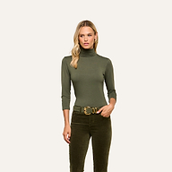 A woman wearing the Aja Turtleneck, a soft knit olive green slim fit top, paired with matching pants and a gold buckle belt stands facing forward against a plain light background.