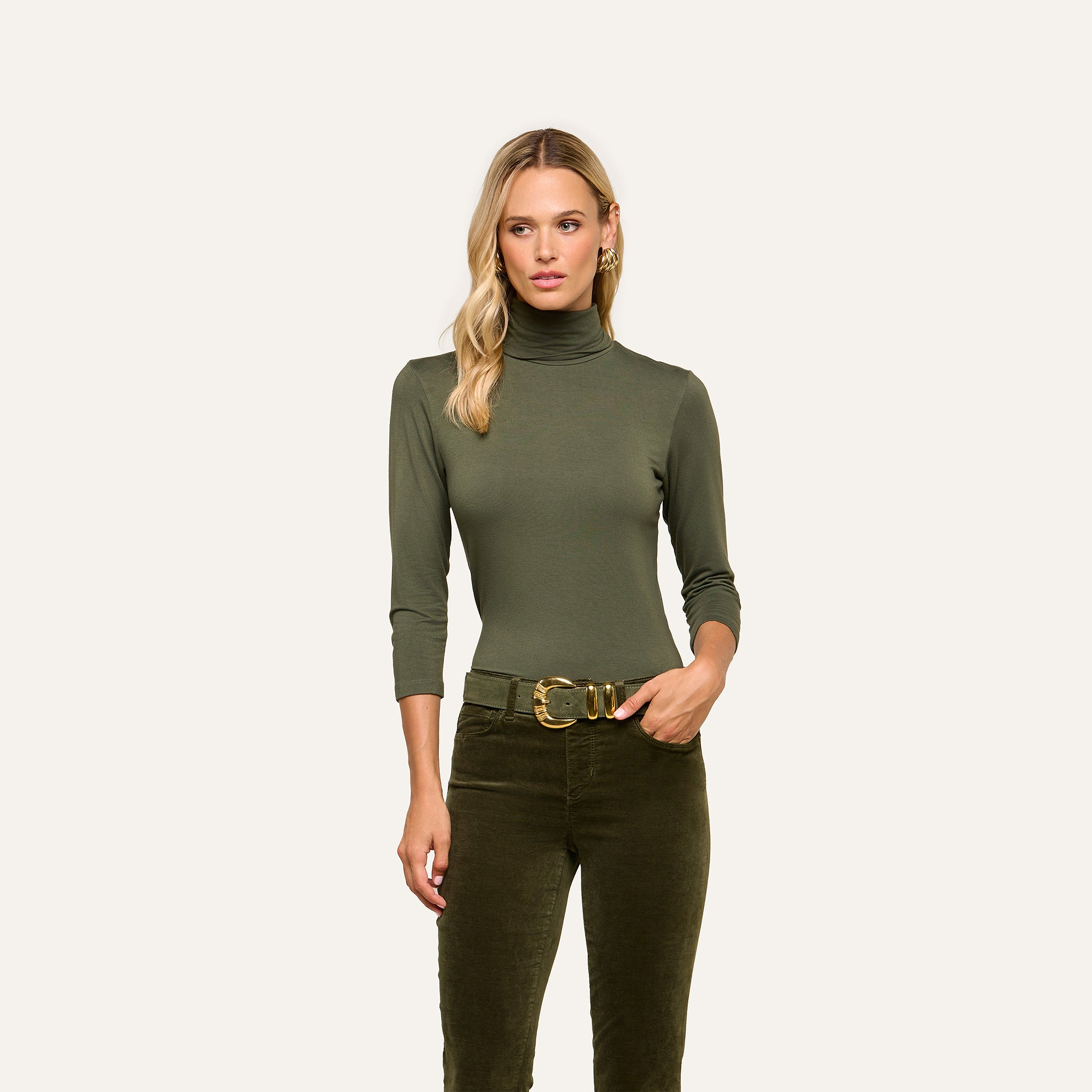 A woman wearing the Aja Turtleneck, a soft knit olive green slim fit top, paired with matching pants and a gold buckle belt stands facing forward against a plain light background.