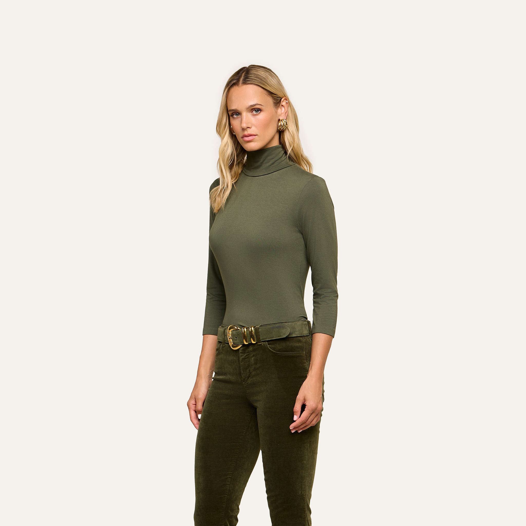 A woman with long blonde hair wears the Aja Turtleneck in soft olive green knit, paired with matching slim fit pants and a wide belt, standing against a plain light background.