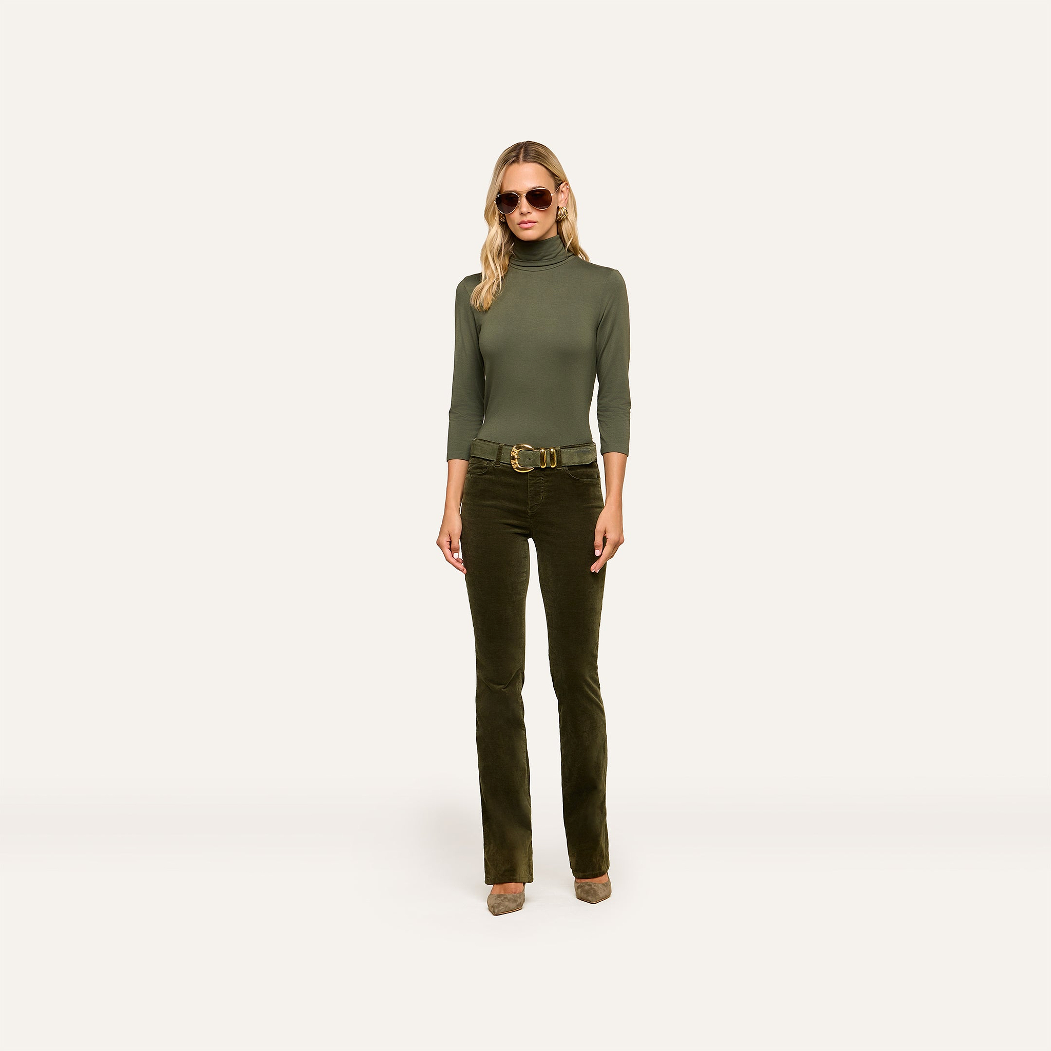 A woman wearing sunglasses, the Aja Turtleneck in a slim fit green style, dark green flared pants with a belt, and heels stands against a plain light background.