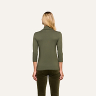 A person with long blonde hair is shown from the back, wearing the Aja Turtleneck—a fitted, olive green top with three-quarter sleeves in soft knit fabric—paired with matching green pants against a plain background. The material accentuates the slim fit silhouette.