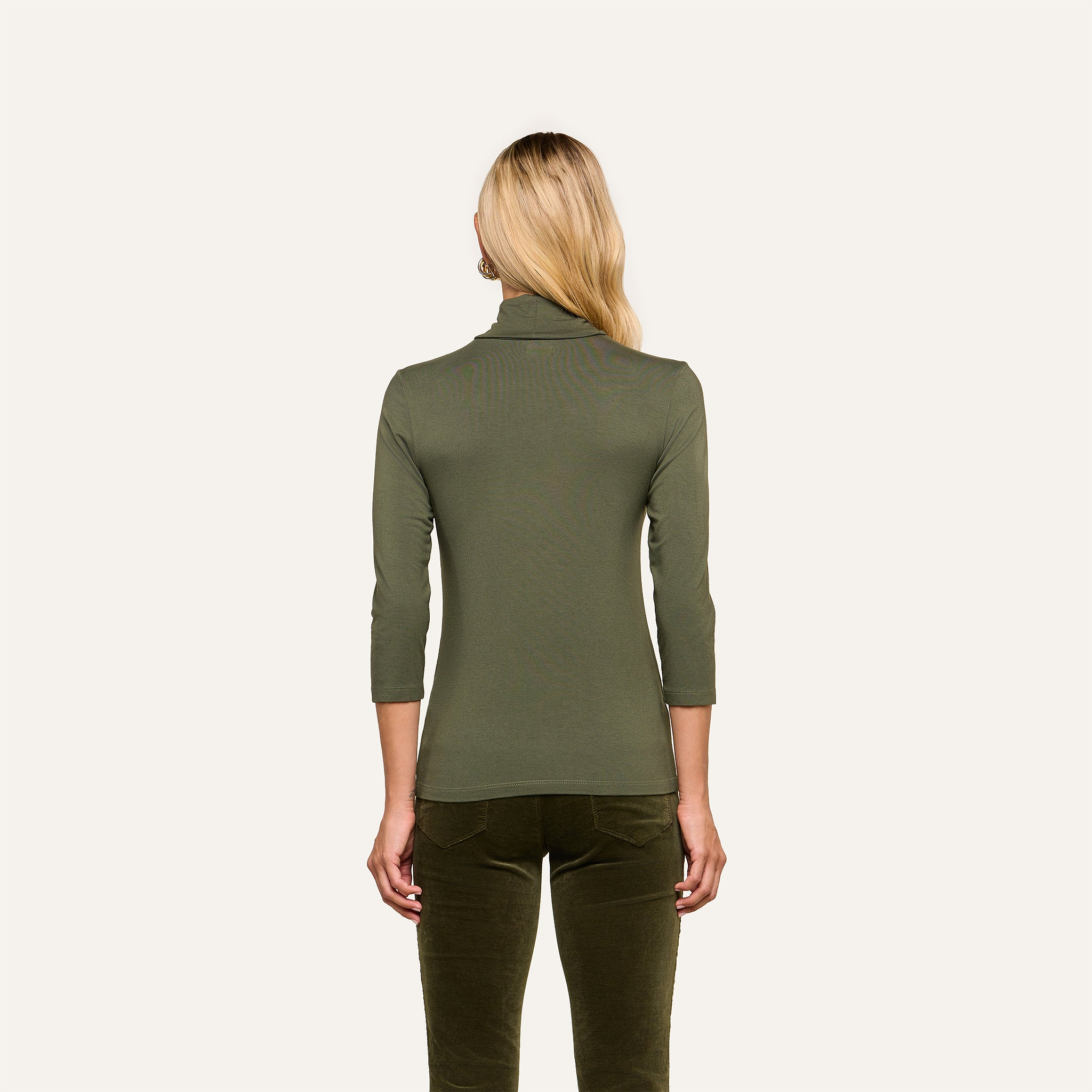 A person with long blonde hair is shown from the back, wearing the Aja Turtleneck—a fitted, olive green top with three-quarter sleeves in soft knit fabric—paired with matching green pants against a plain background. The material accentuates the slim fit silhouette.
