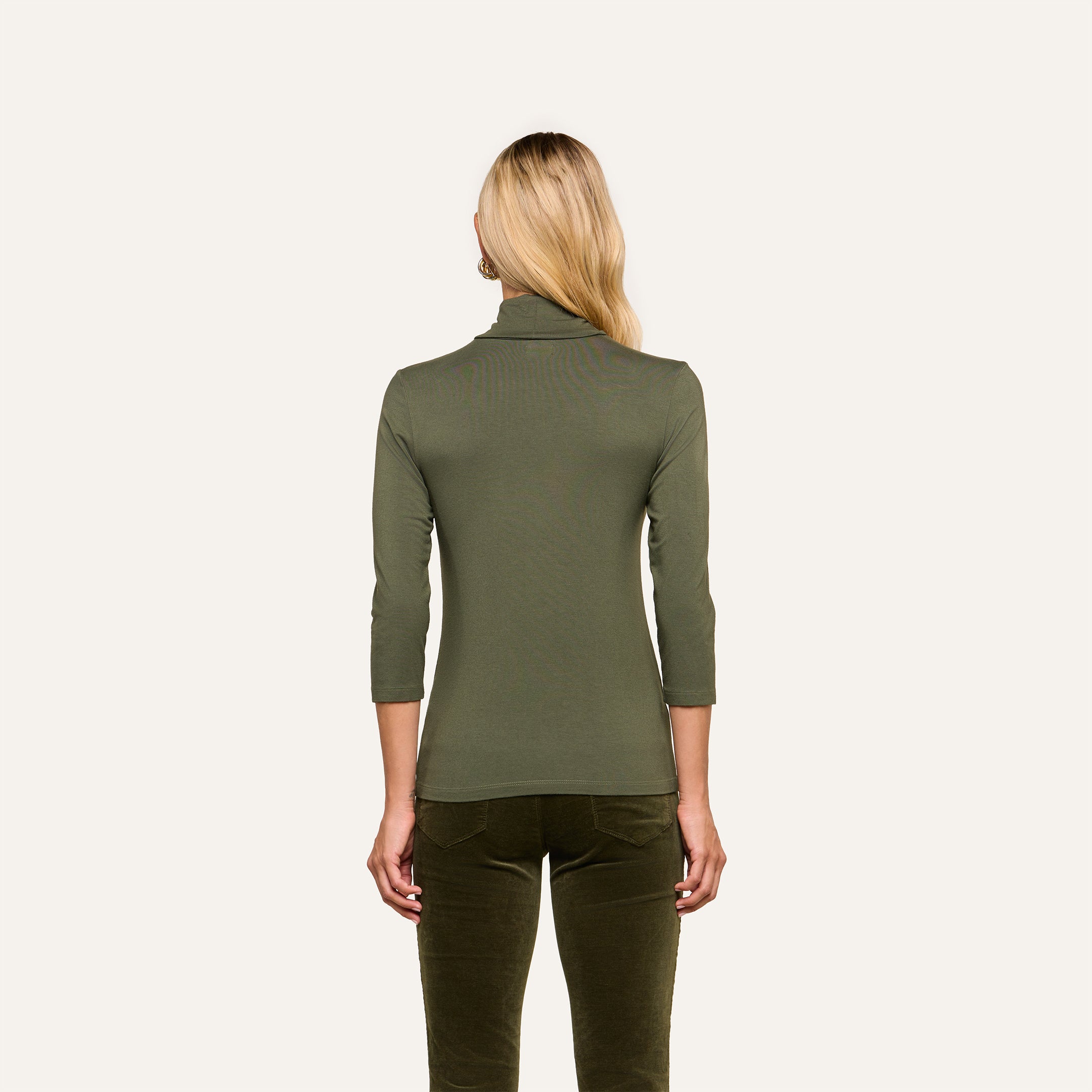 A person with long blonde hair is shown from the back, wearing the Aja Turtleneck—a fitted, olive green top with three-quarter sleeves in soft knit fabric—paired with matching green pants against a plain background. The material accentuates the slim fit silhouette.