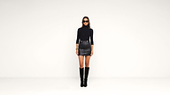 Woman wearing a black turtleneck, black leather mini skirt, knee-high black boots, and sunglasses stands against a plain white background.