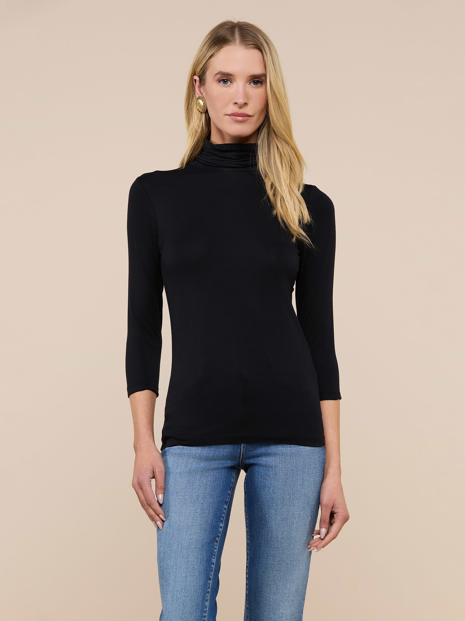 A woman with long blonde hair wears the Aja Turtleneck, a slim-fit black top with three-quarter sleeves, paired with blue jeans and standing against a plain light background.