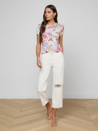 Wearing the bestselling Ressi Fitted Tee in a vibrant print, white wide-leg pants featuring a ripped knee, and beige strappy sandals, the woman stands against a plain white background.