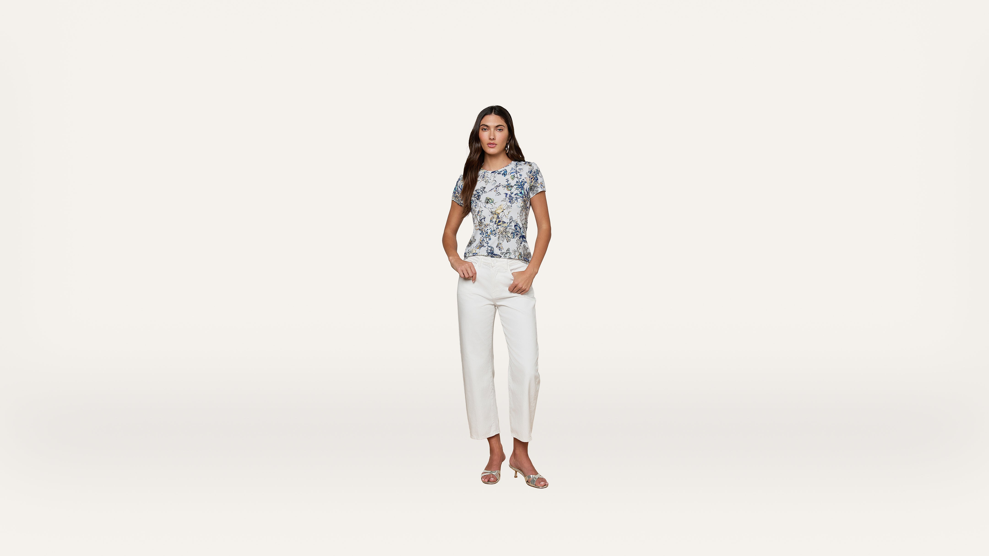 Woman standing against a plain white background, wearing a blue and white patterned short-sleeve top, white pants, and open-toe sandals, with her hands in her pockets.