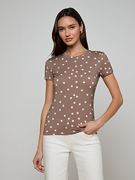 A woman stands against a light background, wearing the Ressi Fitted Tee—a slim cut brown short-sleeve shirt with white polka dots—paired with white pants, with one hand in her pocket.
