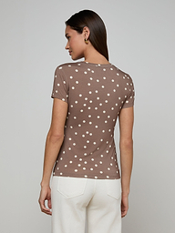 A woman stands facing away, wearing the Ressi Fitted Tee—a slim cut brown short-sleeve shirt with white polka dots—and white pants, set against a plain light background.