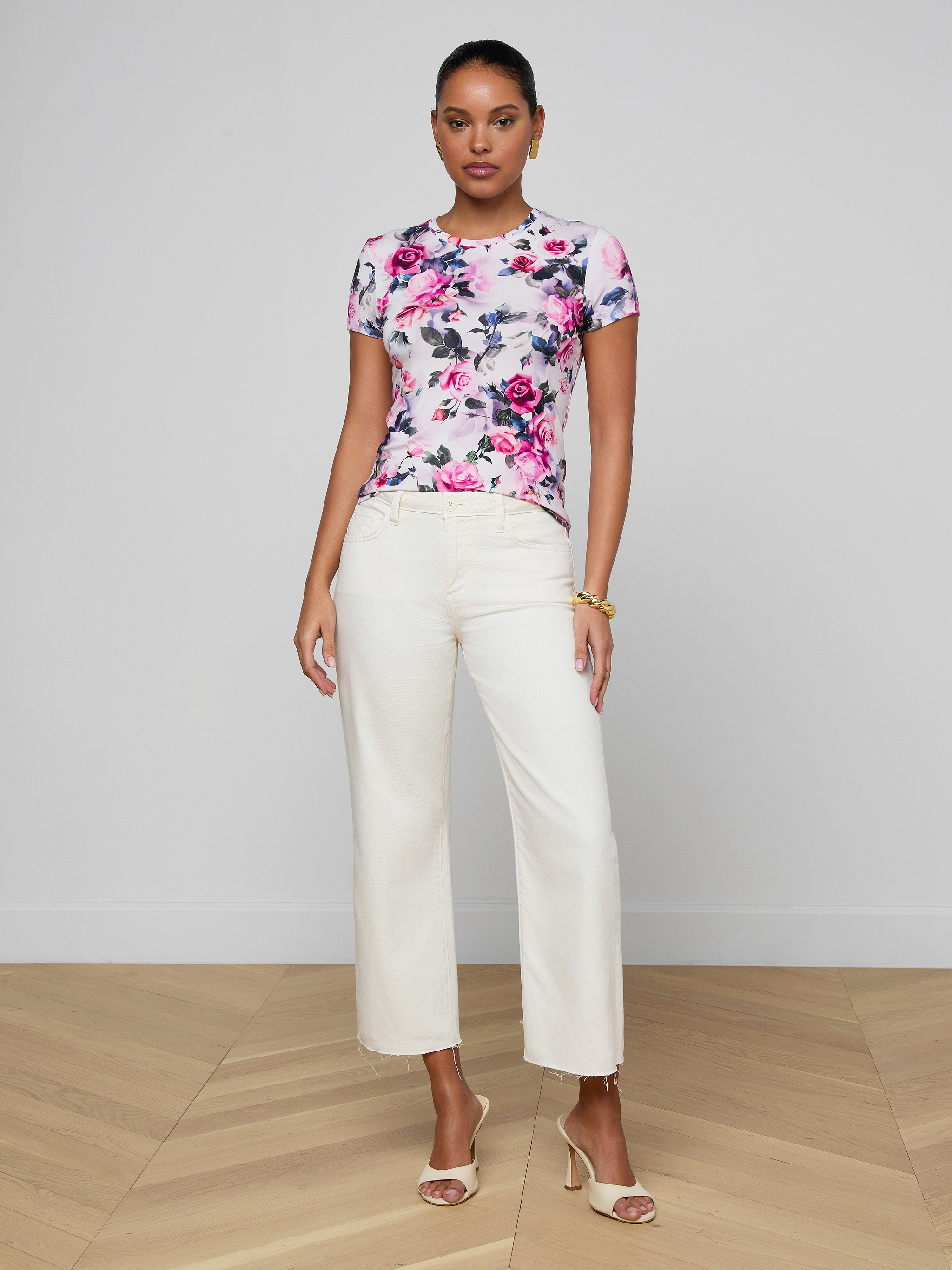 A woman stands against a white background, wearing the Ressi Fitted Tee in a short-sleeve pink rose print stretch jersey knit, paired with white wide-leg pants, nude high-heeled sandals, and gold bracelets.