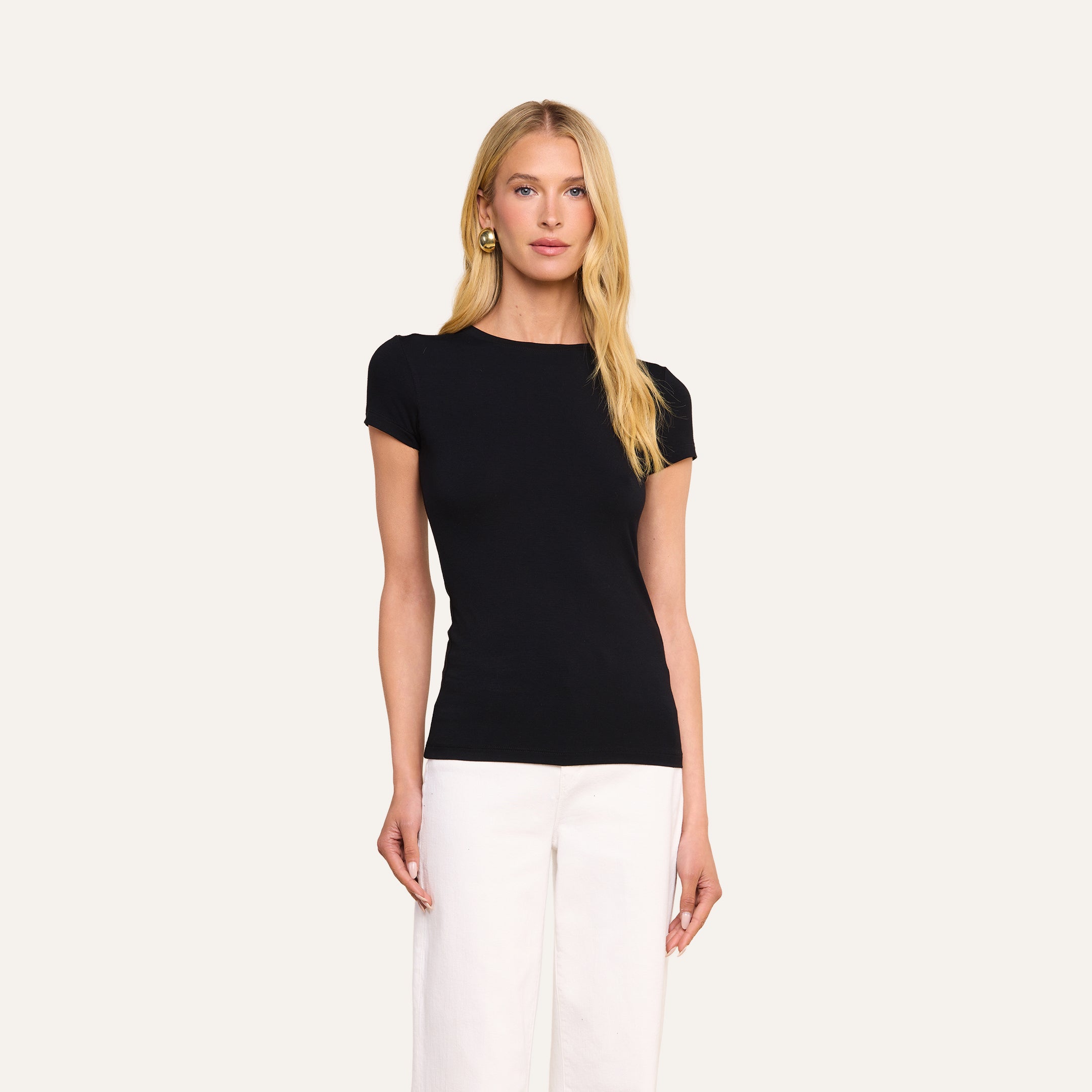 A woman with long blonde hair wears the form-fitting black Ressi Fitted Tee and white pants, standing against a plain light background.