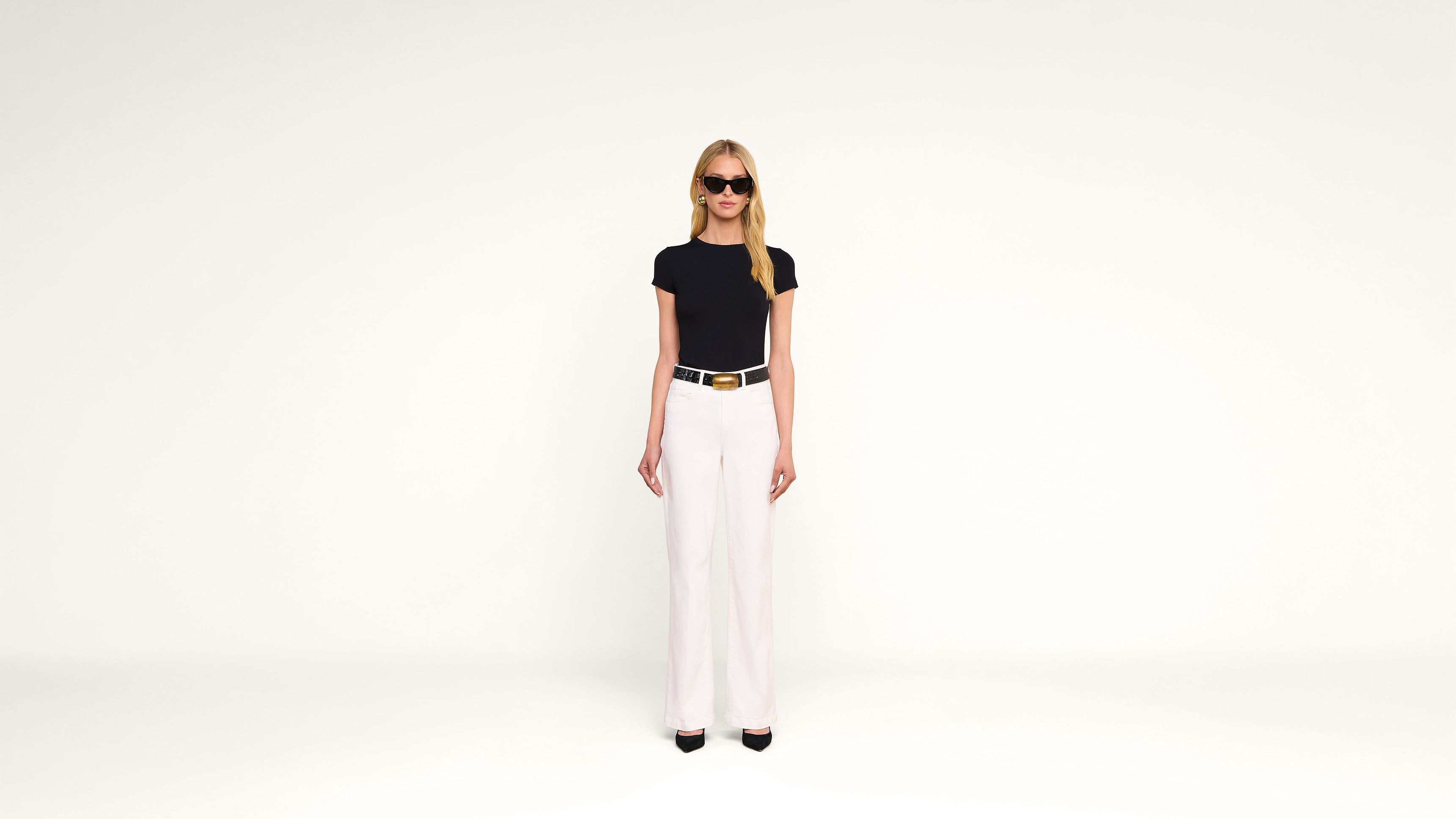 A woman wearing sunglasses, a black t-shirt, white high-waisted pants, and black heels stands against a plain white background, facing forward.