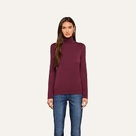 A woman with long brown hair wears the Lani Turtleneck in maroon and blue jeans, standing against a plain off-white background and looking straight at the camera with a neutral expression.