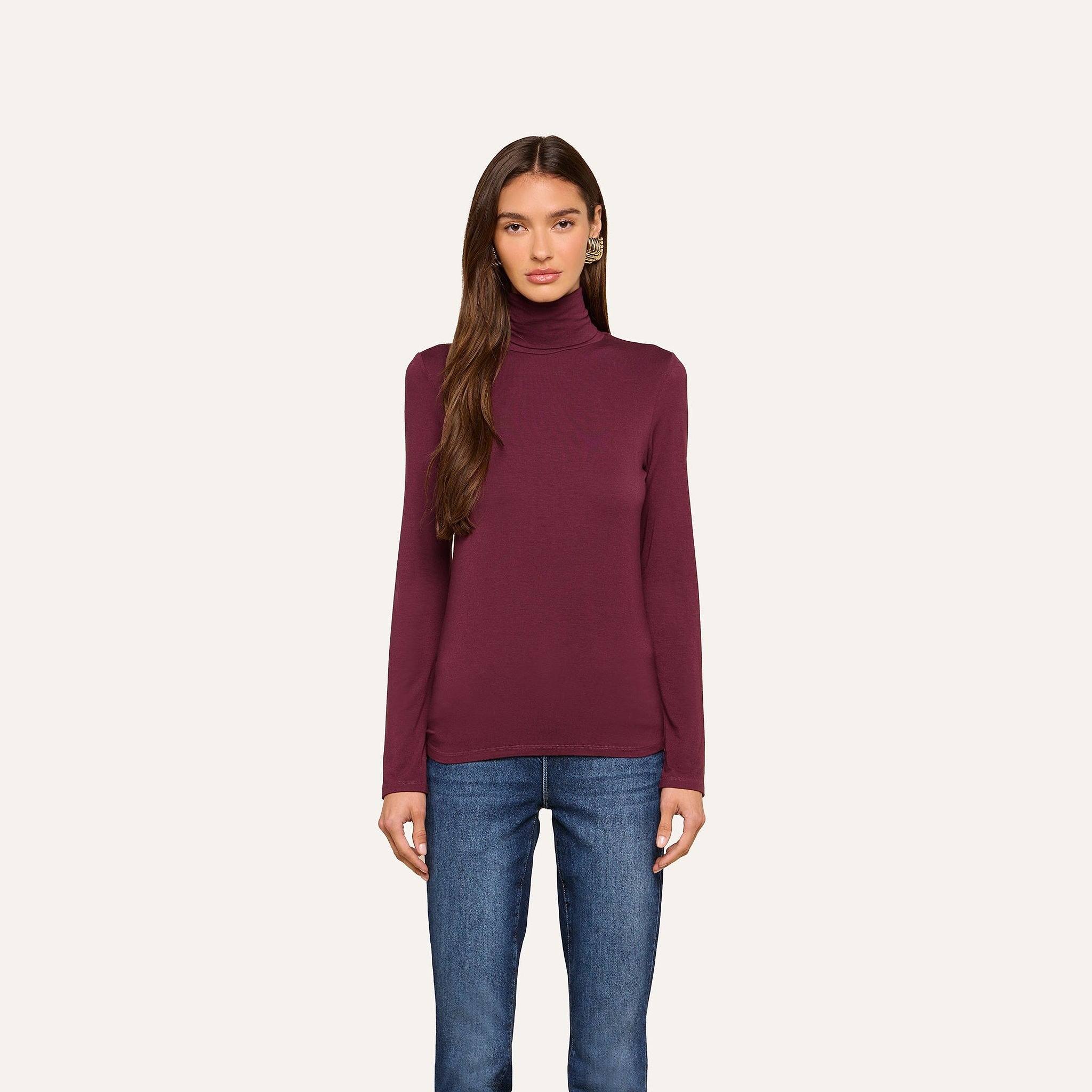 A woman with long brown hair wears the Lani Turtleneck in maroon and blue jeans, standing against a plain off-white background and looking straight at the camera with a neutral expression.