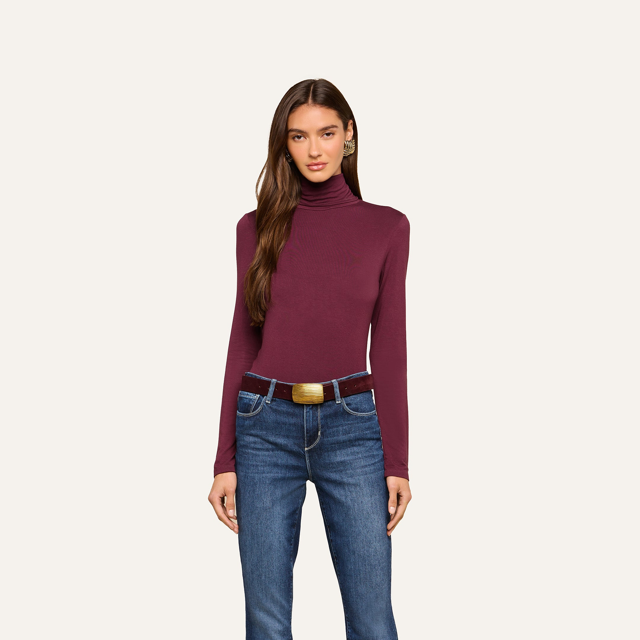 A woman is wearing the Lani Turtleneck, a maroon slim-fit long-sleeve top, tucked into blue jeans with a brown belt, standing against a plain light background.