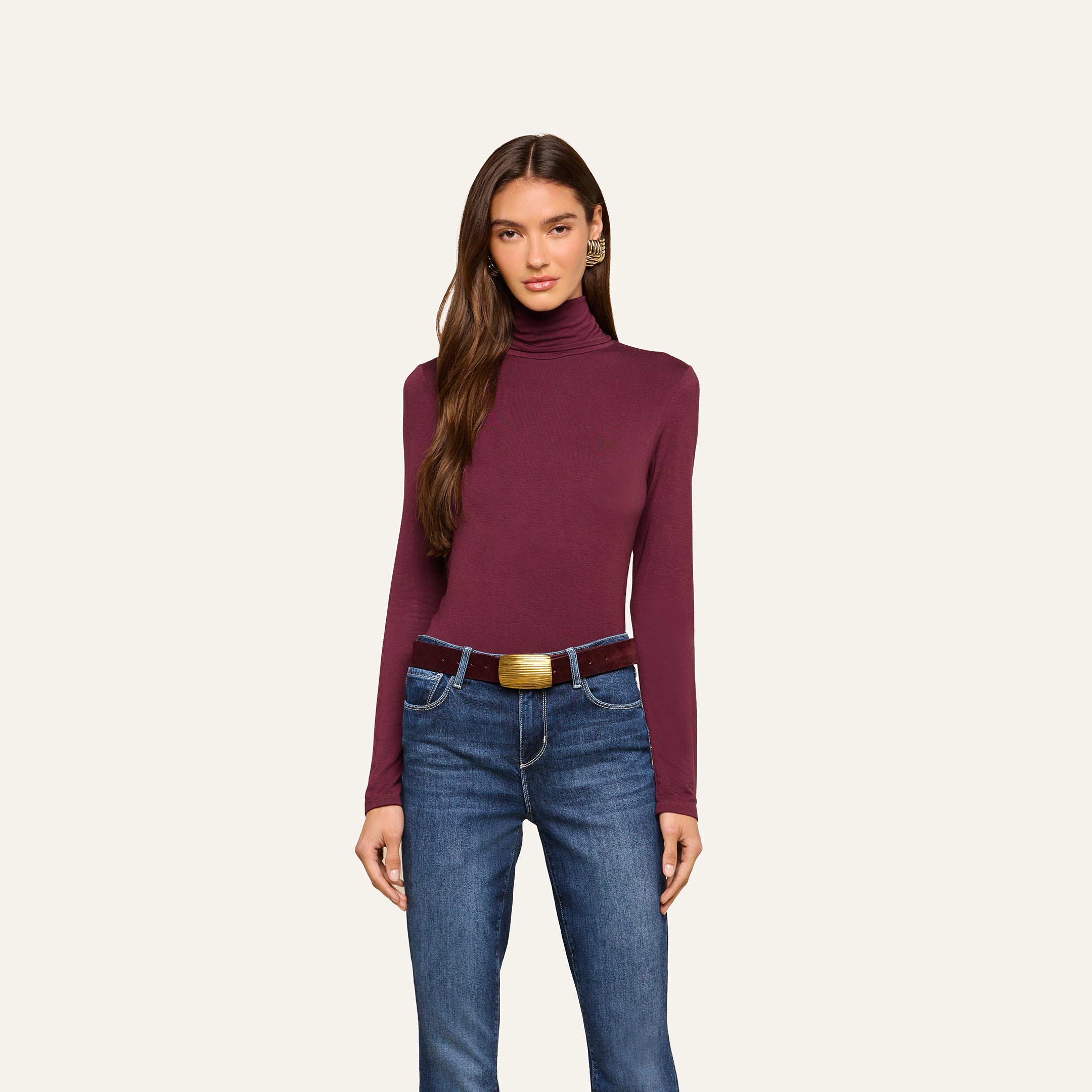 A woman is wearing the Lani Turtleneck, a maroon slim-fit long-sleeve top, tucked into blue jeans with a brown belt, standing against a plain light background.