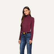 A woman wears the Lani Turtleneck in maroon, paired with blue jeans and a brown belt, standing against a plain light background. She faces forward, with one hand by her side and the other resting on her leg—a classic look perfect for Fall 2026.