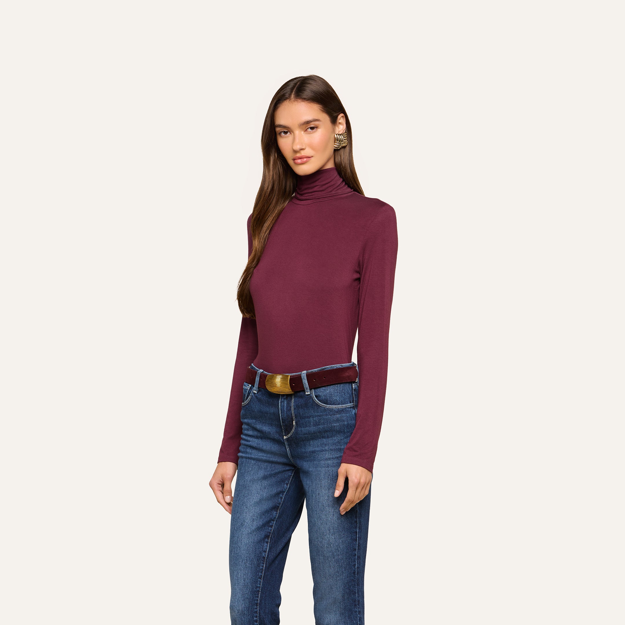 A woman wears the Lani Turtleneck in maroon, paired with blue jeans and a brown belt, standing against a plain light background. She faces forward, with one hand by her side and the other resting on her leg—a classic look perfect for Fall 2026.