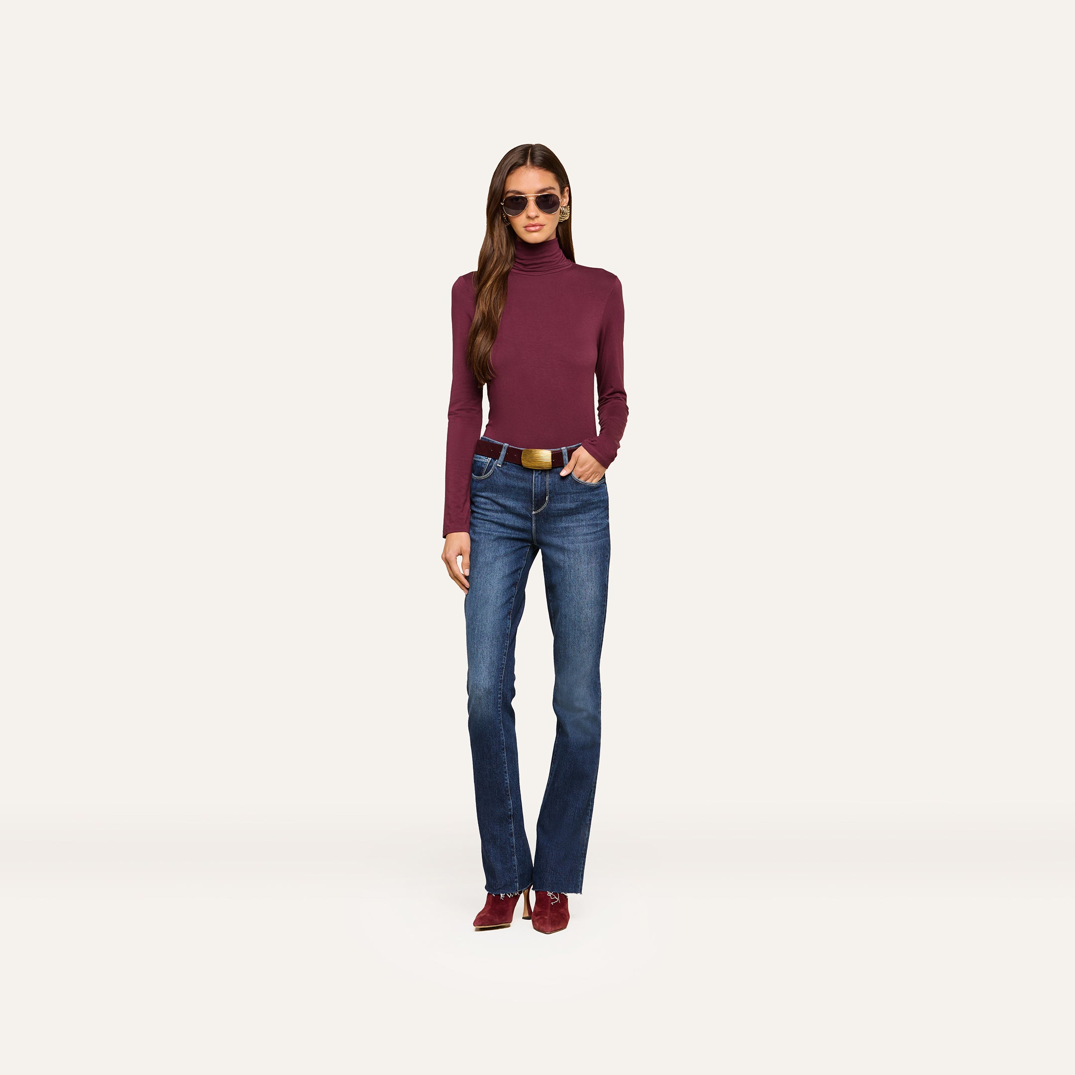 A woman wearing sunglasses, the Lani Turtleneck in a slim fit burgundy shade, blue jeans with a belt, and matching burgundy shoes stands facing forward against a plain light background.