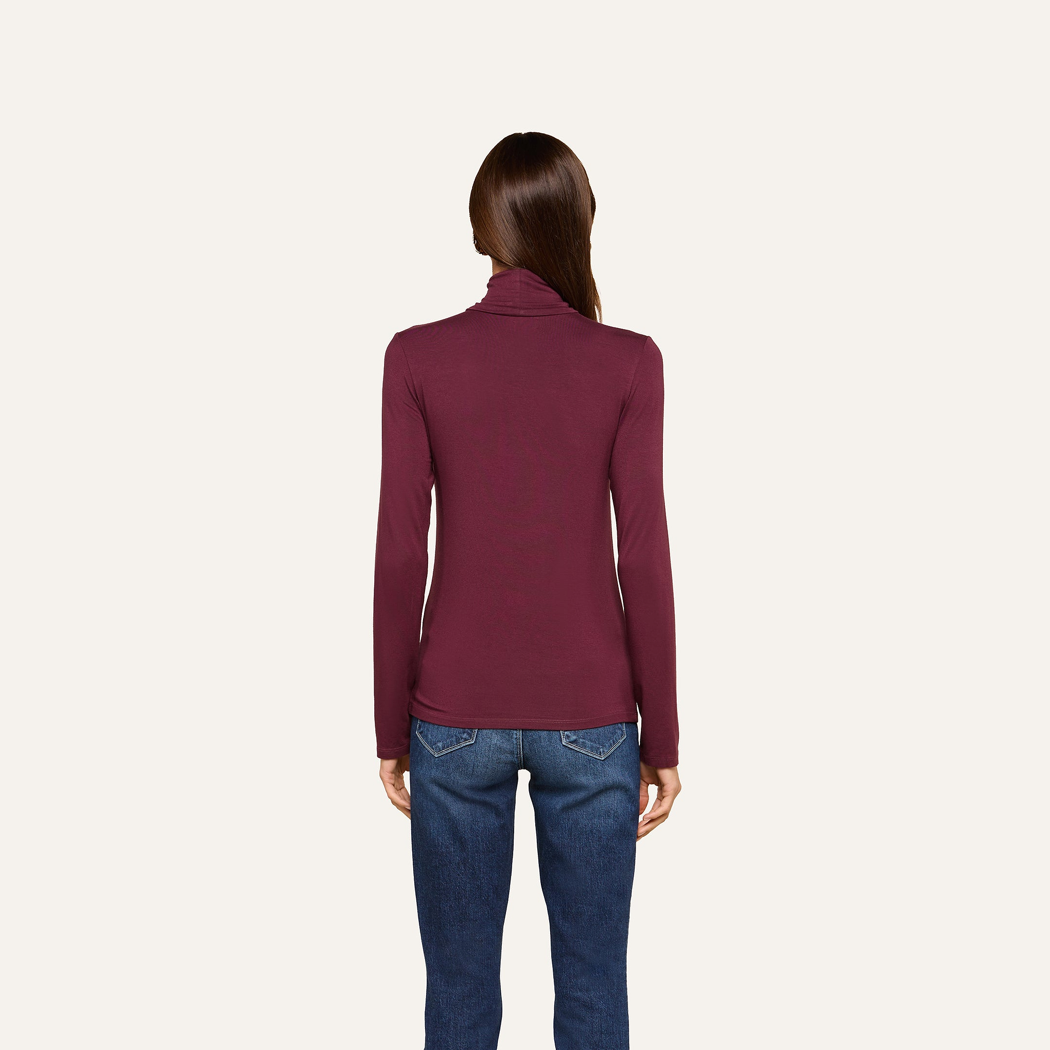 A woman stands facing away, wearing the slim fit Lani Turtleneck in maroon paired with blue jeans against a plain, light background—a perfect look for Fall 2026.
