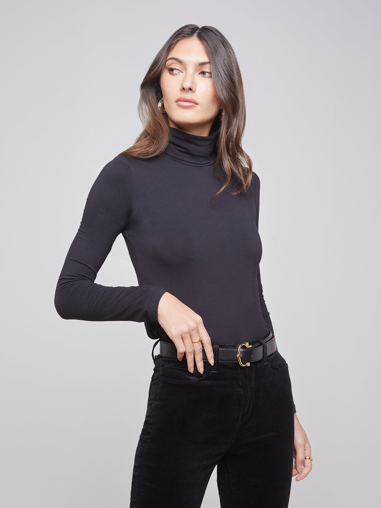 Wearing the Lani Turtleneck and black belted pants, a woman stands against a plain light background, looking to the side with one hand on her hip.