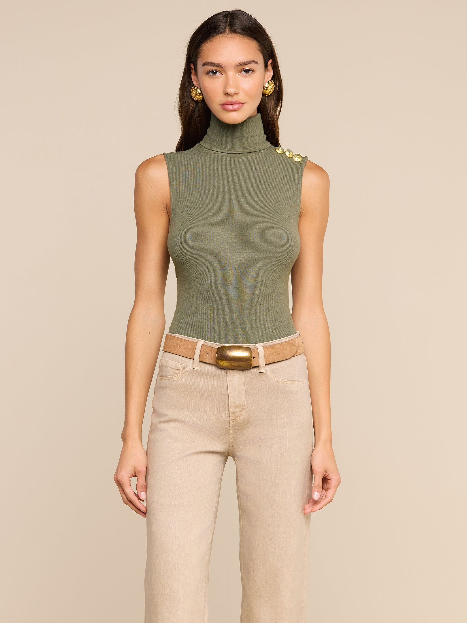 A woman models the Ceci Sleeveless Turtleneck in ash olive green, styled with beige high-waisted pants, a wide brown belt, and gold earrings, standing against a plain light background.