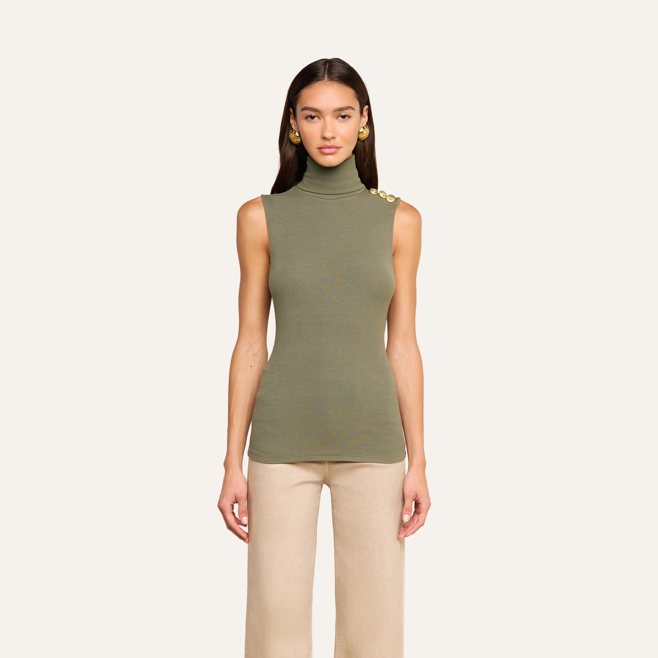 A woman models the Ceci Sleeveless Turtleneck in ash olive green—a ribbed knit top with gold shoulder buttons—paired with beige pants, standing against a plain light background and facing forward.