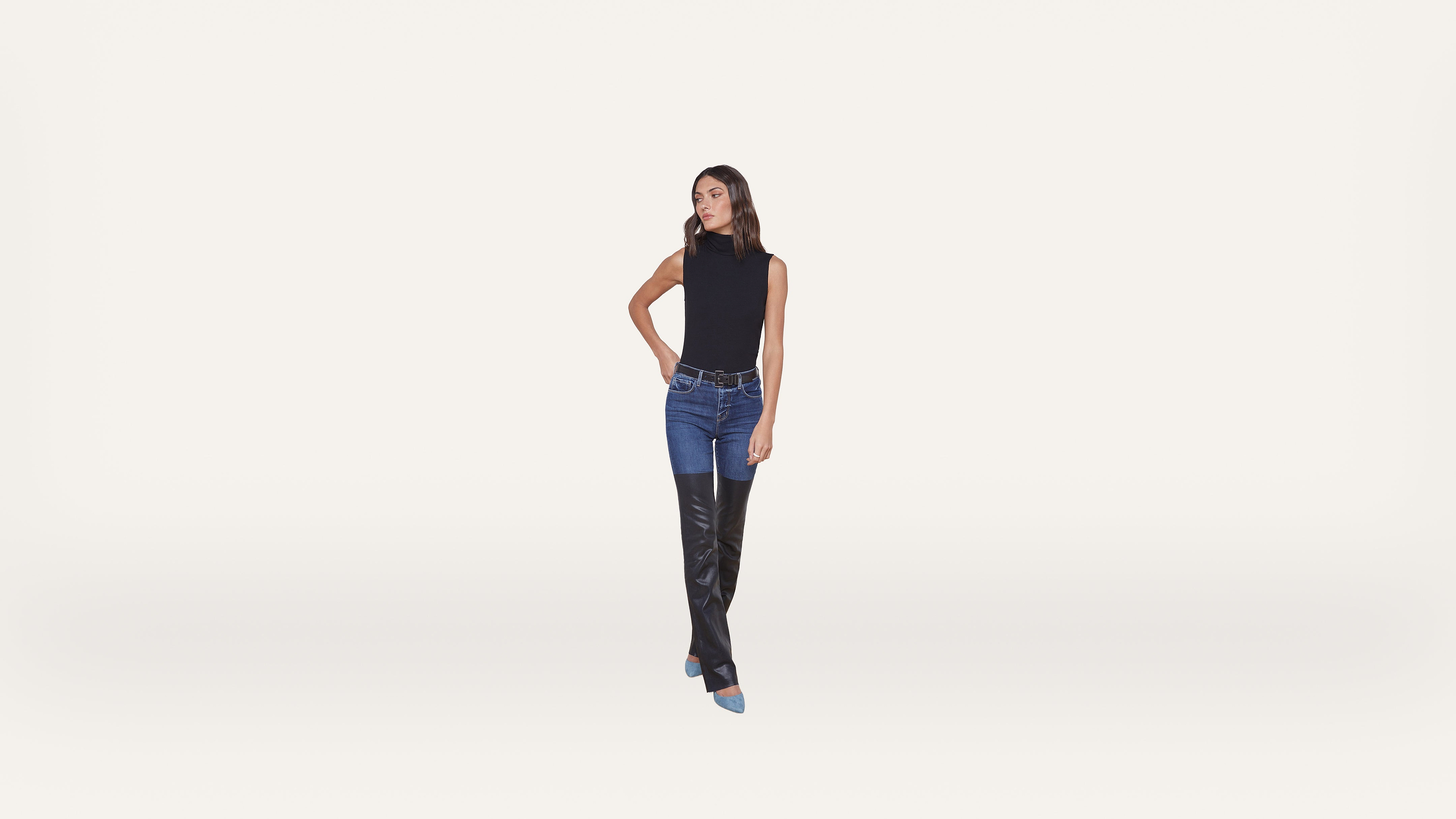 Woman standing against a plain white background, wearing a sleeveless black top, blue jeans, and black over-the-knee boots with blue pointed toes. She has straight brown hair and her left hand is on her hip.