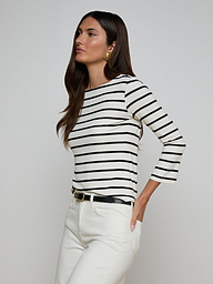 Wearing the Lucille Striped Boatneck Top—a black and white, long-sleeve shirt featuring a boat neckline—paired with white pants and a black belt, the woman stands with one hand in her pocket against a plain background. The timeless stripes of the top add classic appeal to her look.