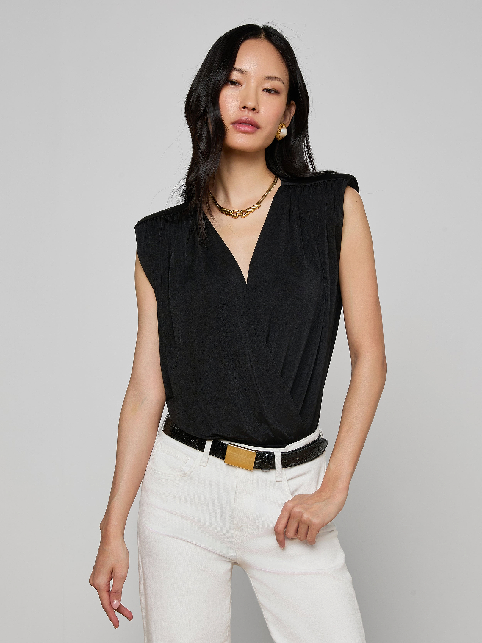 Woman wearing the Akari Bodysuit—a sleeveless black top with subtle shoulder pads—paired with white pants, a black belt with a gold buckle, gold necklace, and earrings, standing against an off-white background.