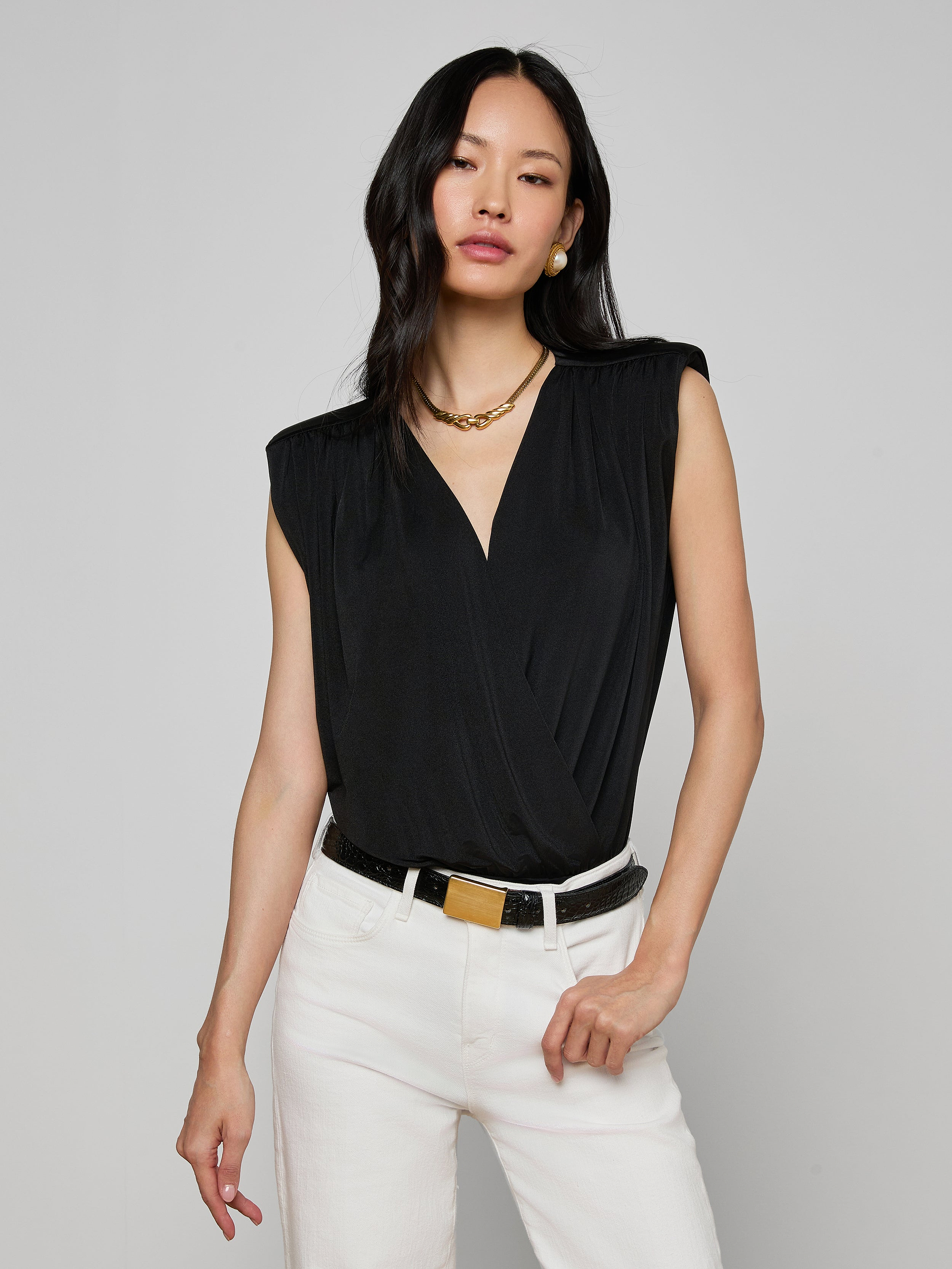 Woman wearing the Akari Bodysuit—a sleeveless black top with subtle shoulder pads—paired with white pants, a black belt with a gold buckle, gold necklace, and earrings, standing against an off-white background.