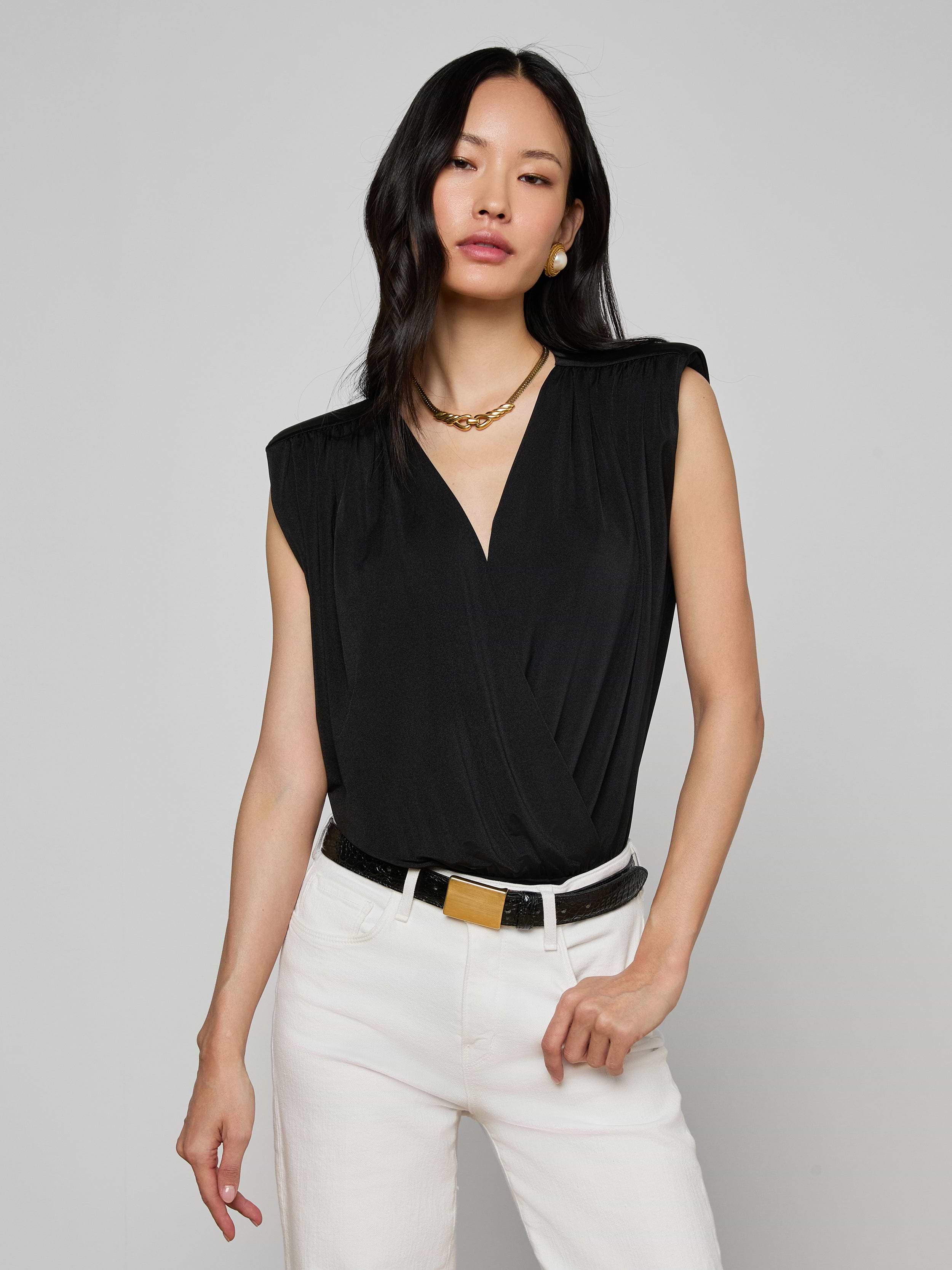 Woman wearing the Akari Bodysuit—a sleeveless black top with subtle shoulder pads—paired with white pants, a black belt with a gold buckle, gold necklace, and earrings, standing against an off-white background.