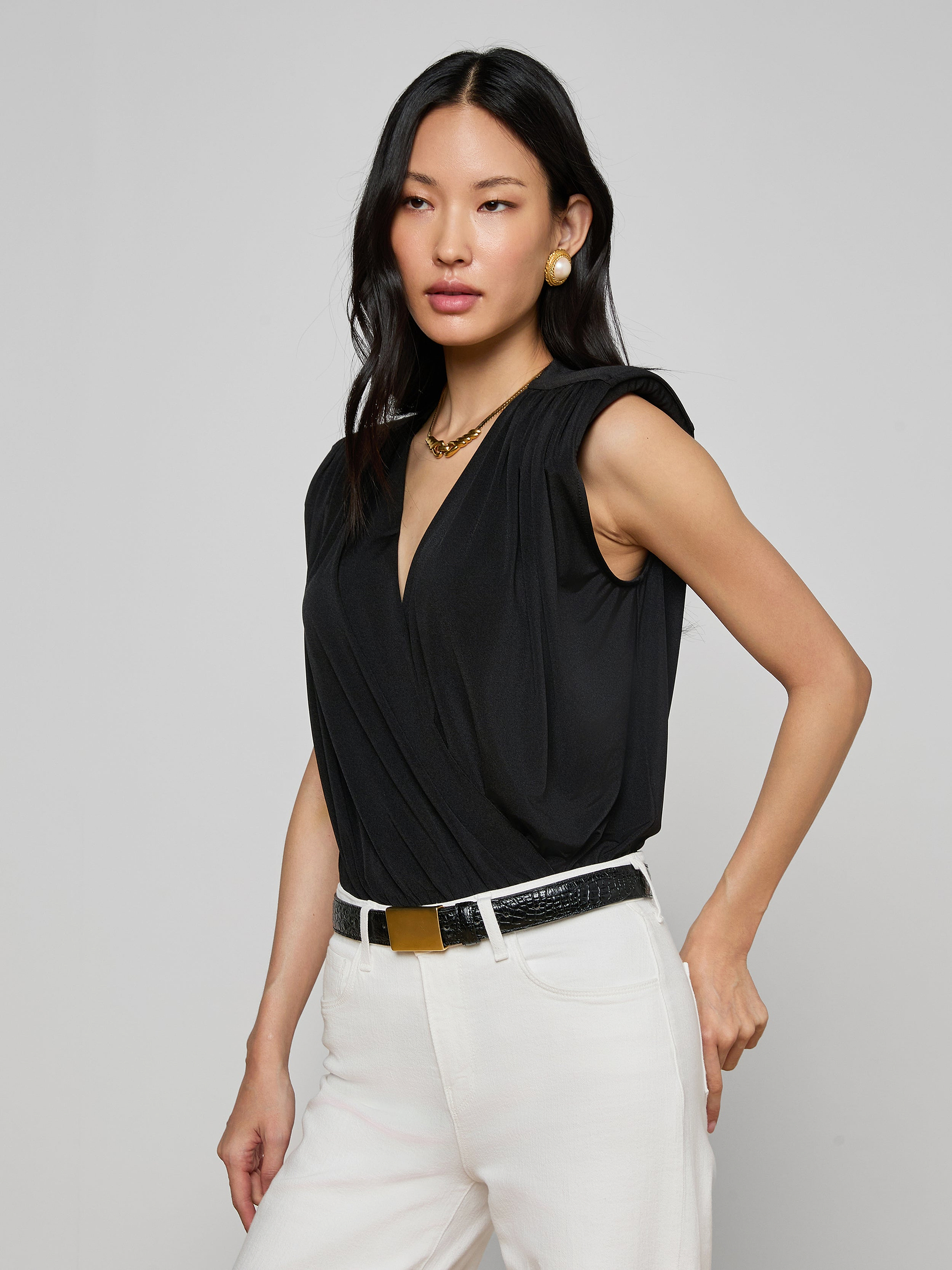 A woman with long black hair wears the Akari Bodysuit—a sleeveless black liquid jersey top with a deep V-neck—paired with white pants. She accessorizes her look with gold earrings, a necklace, and a black belt featuring a gold buckle while posing against a plain light background.