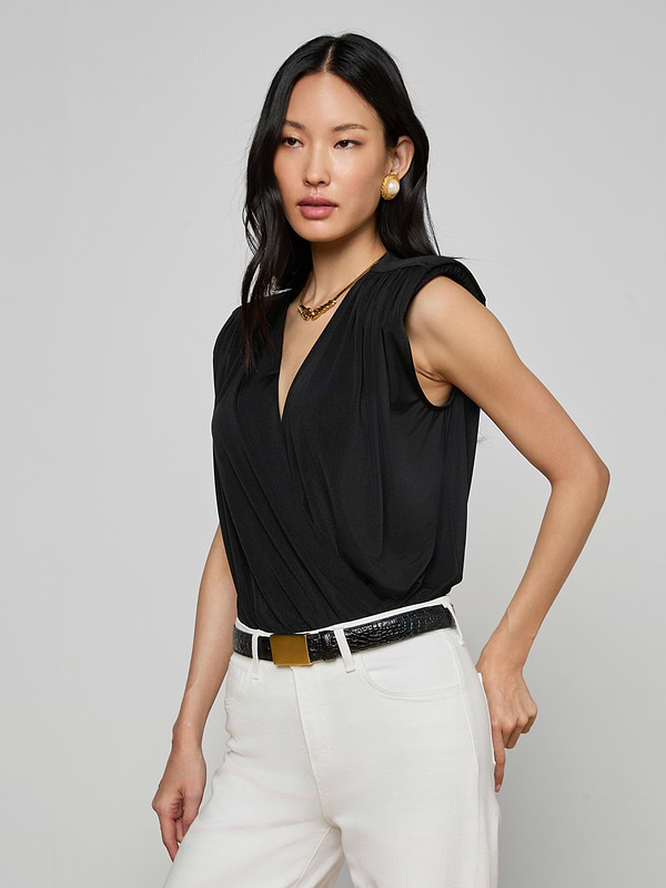 A woman with long black hair wears the Akari Bodysuit—a sleeveless black liquid jersey top with a deep V-neck—paired with white pants. She accessorizes her look with gold earrings, a necklace, and a black belt featuring a gold buckle while posing against a plain light background.