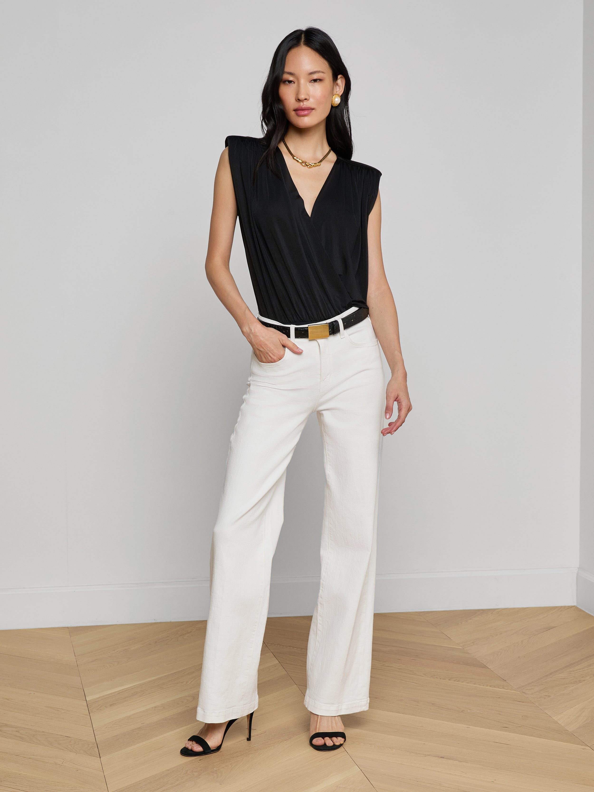 A woman stands against a plain background in a sleeveless black Akari Bodysuit, paired with white wide-leg pants, a black belt, black heels, and a gold necklace. With one hand in her pocket, she poses in a relaxed manner.