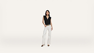 A woman stands against a plain white background, wearing a sleeveless black top, white wide-leg pants, and black heeled sandals, with one hand in her pocket.
