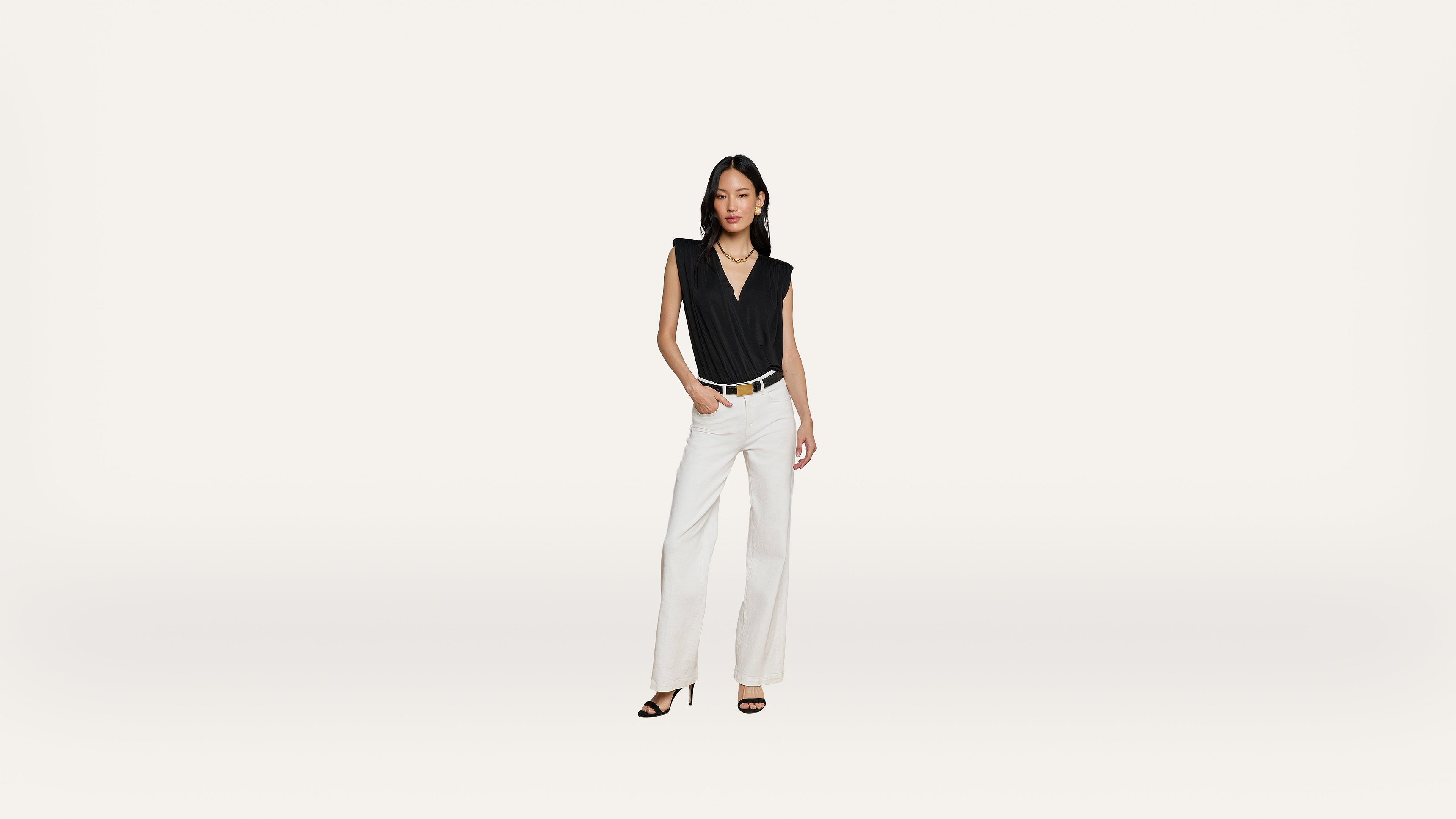 A woman stands against a plain white background, wearing a sleeveless black top, white wide-leg pants, and black heeled sandals, with one hand in her pocket.