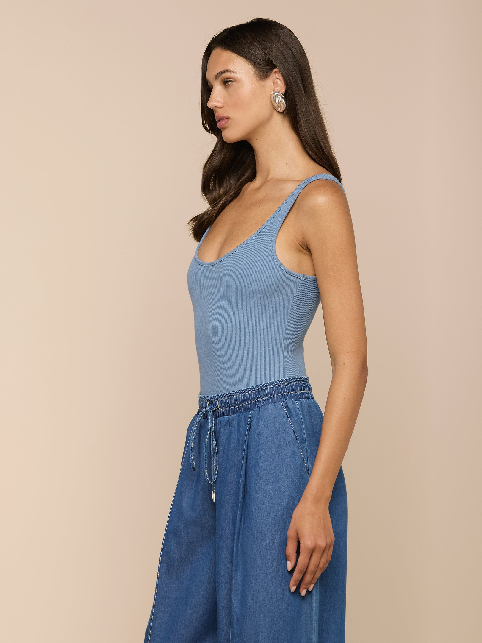 Eine Frau mit langen braunen Haaren, im Profil vor einem beigen Hintergrund, trägt einen blauen „Nica Scoopneck Bodysuit“ mit figurbetonter Passform, eine blaue Hose mit weitem Bein und große silberne Ohrringe.
