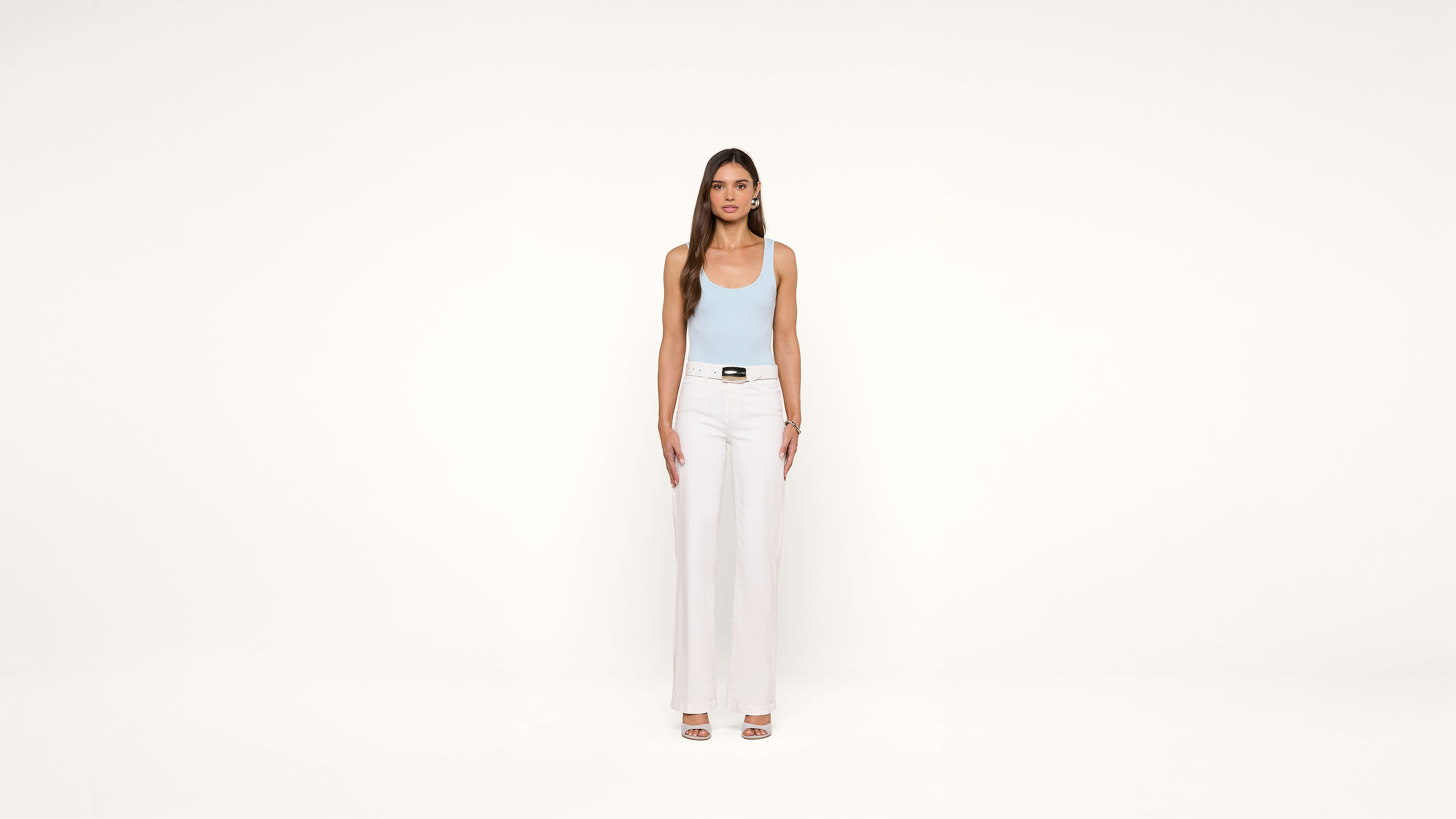 A woman stands facing forward against a plain white background, wearing a light blue tank top, white high-waisted pants, and open-toe heels. Her arms are relaxed by her sides.