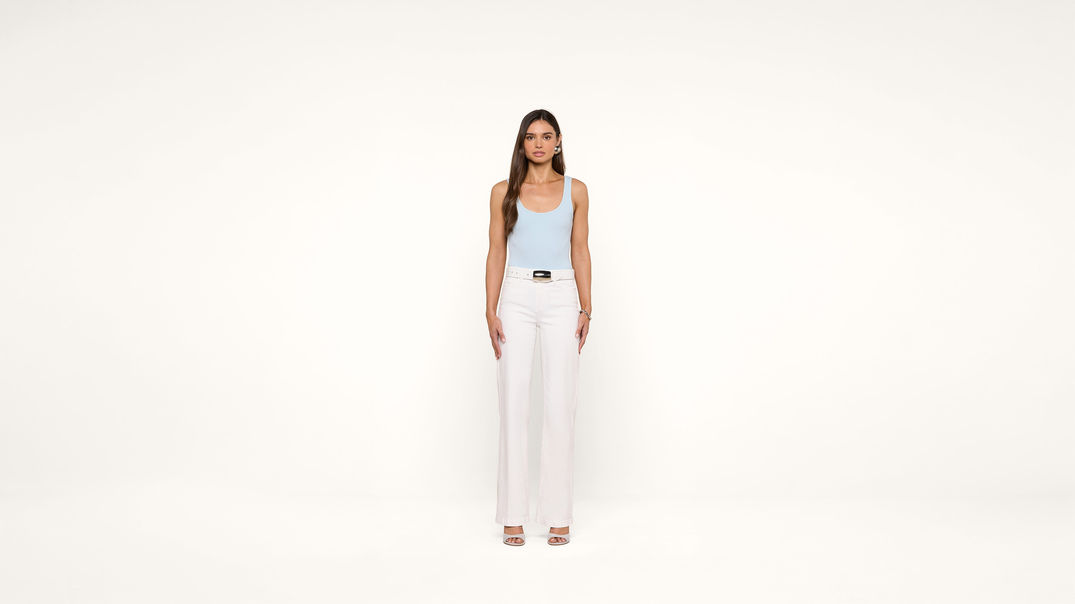 A woman stands facing forward against a plain white background, wearing a light blue tank top, white high-waisted pants, and open-toe heels. Her arms are relaxed by her sides.