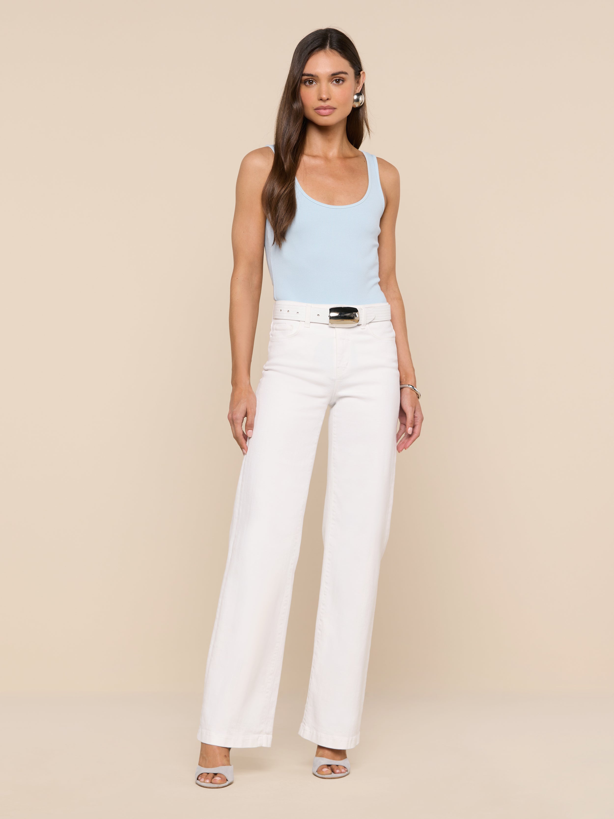 A woman with long brown hair and a neutral expression stands against a plain white background, wearing the Nica Scoopneck Bodysuit in light blue, paired with high-waisted white wide-leg pants, a white belt, and open-toe heels.