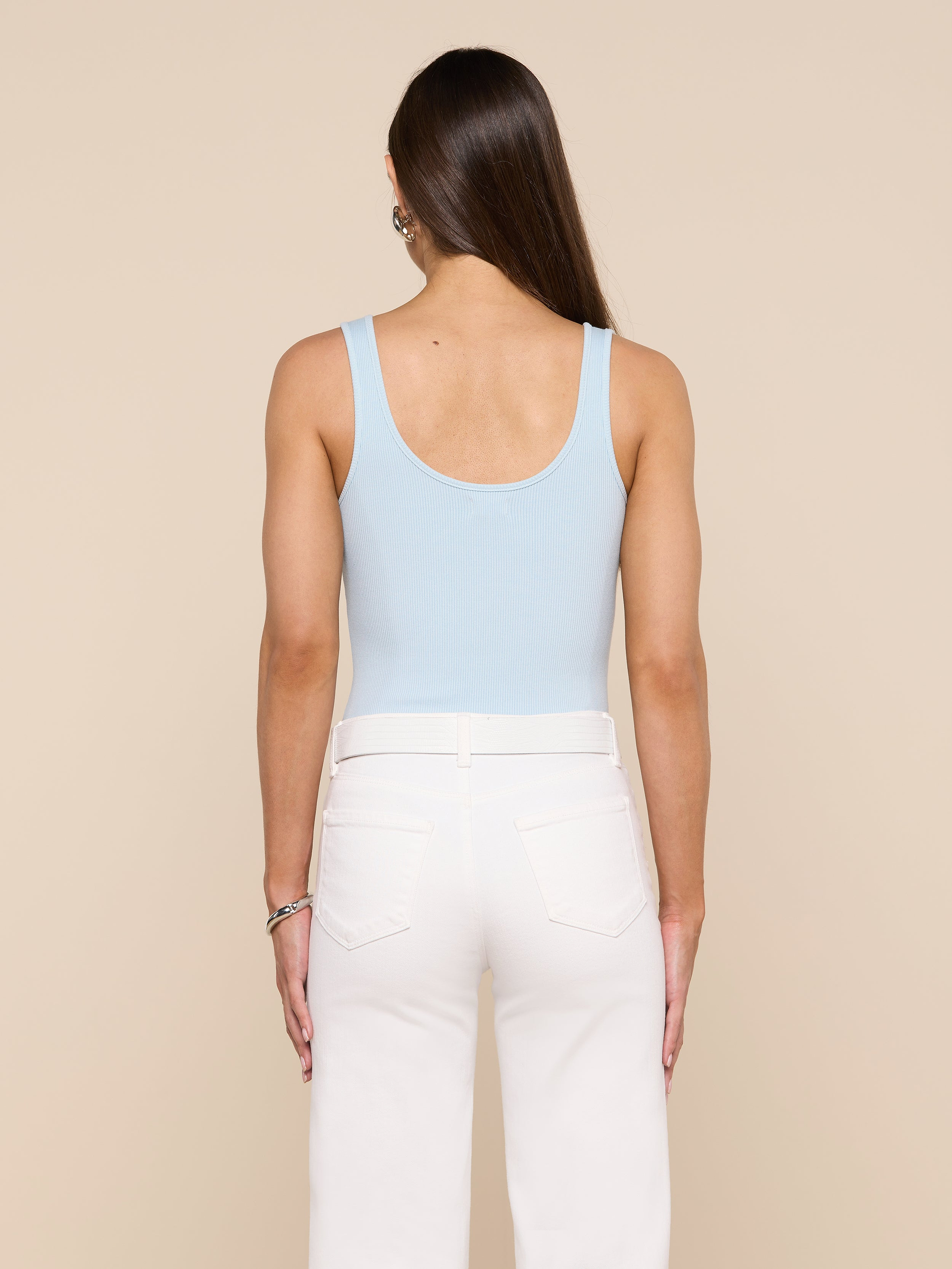 A woman with long brown hair, wearing the Nica Scoopneck Bodysuit in light blue and high-waisted white pants, stands facing away from the camera against a plain light background. The sculpted fit of the bodysuit highlights her silhouette.