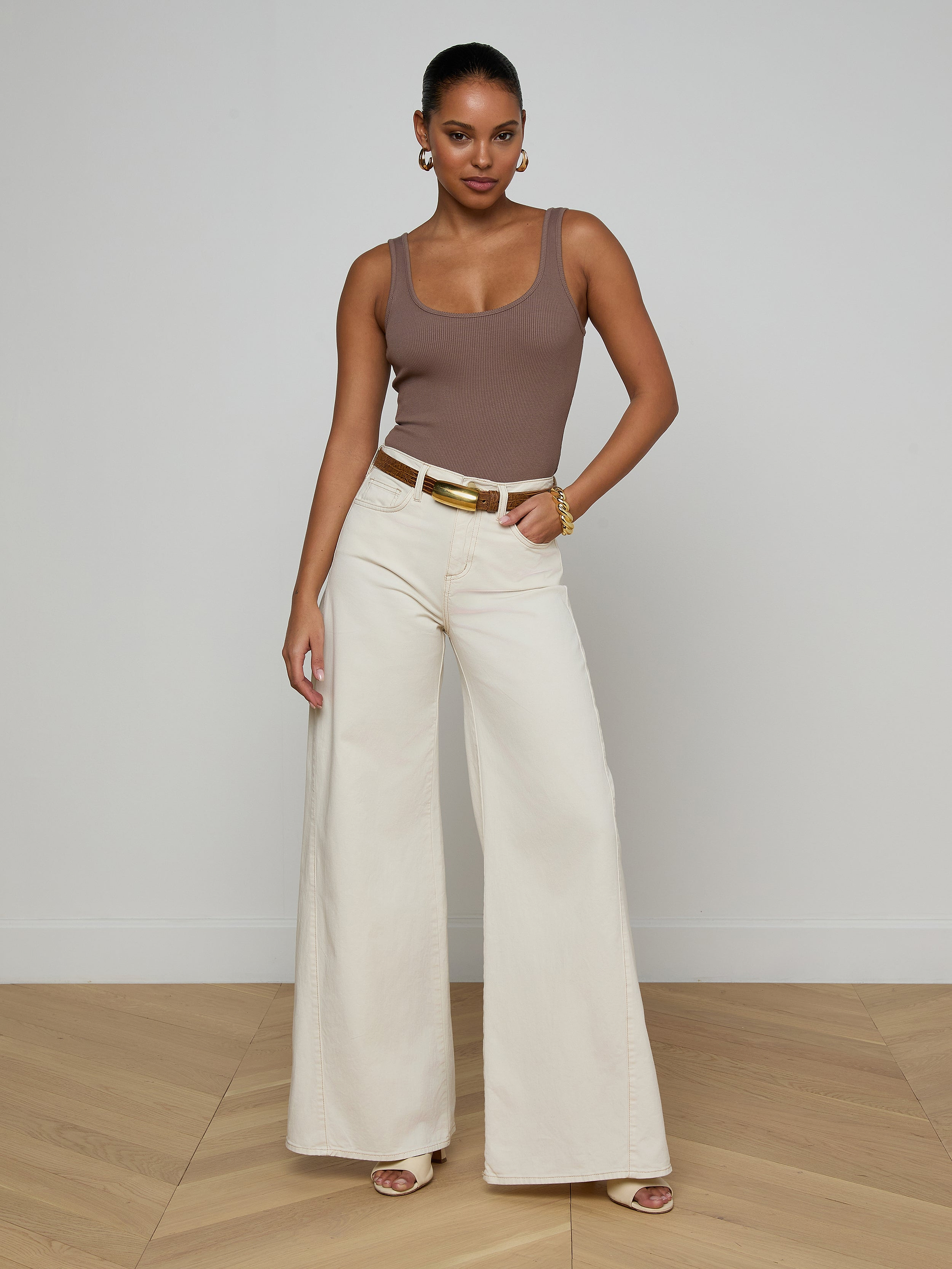 A woman wearing sunglasses, the Nica Scoopneck Bodysuit in taupe with a stretch rib and sculpted fit, blue cropped jeans, and nude heels stands against a plain white background.