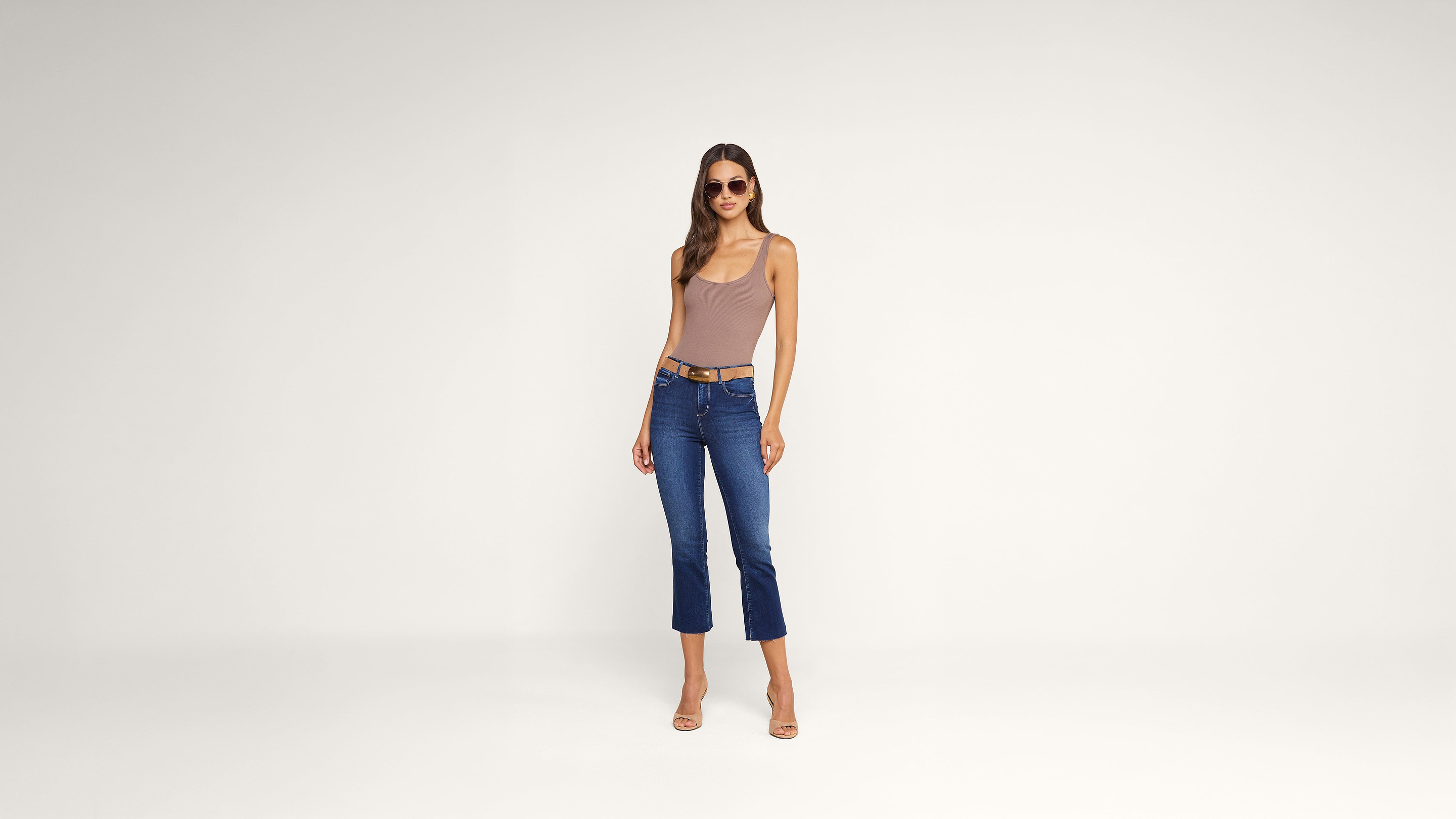 Woman wearing sunglasses, a sleeveless mauve bodysuit, blue cropped jeans with a belt, and nude heels stands against a plain white background.