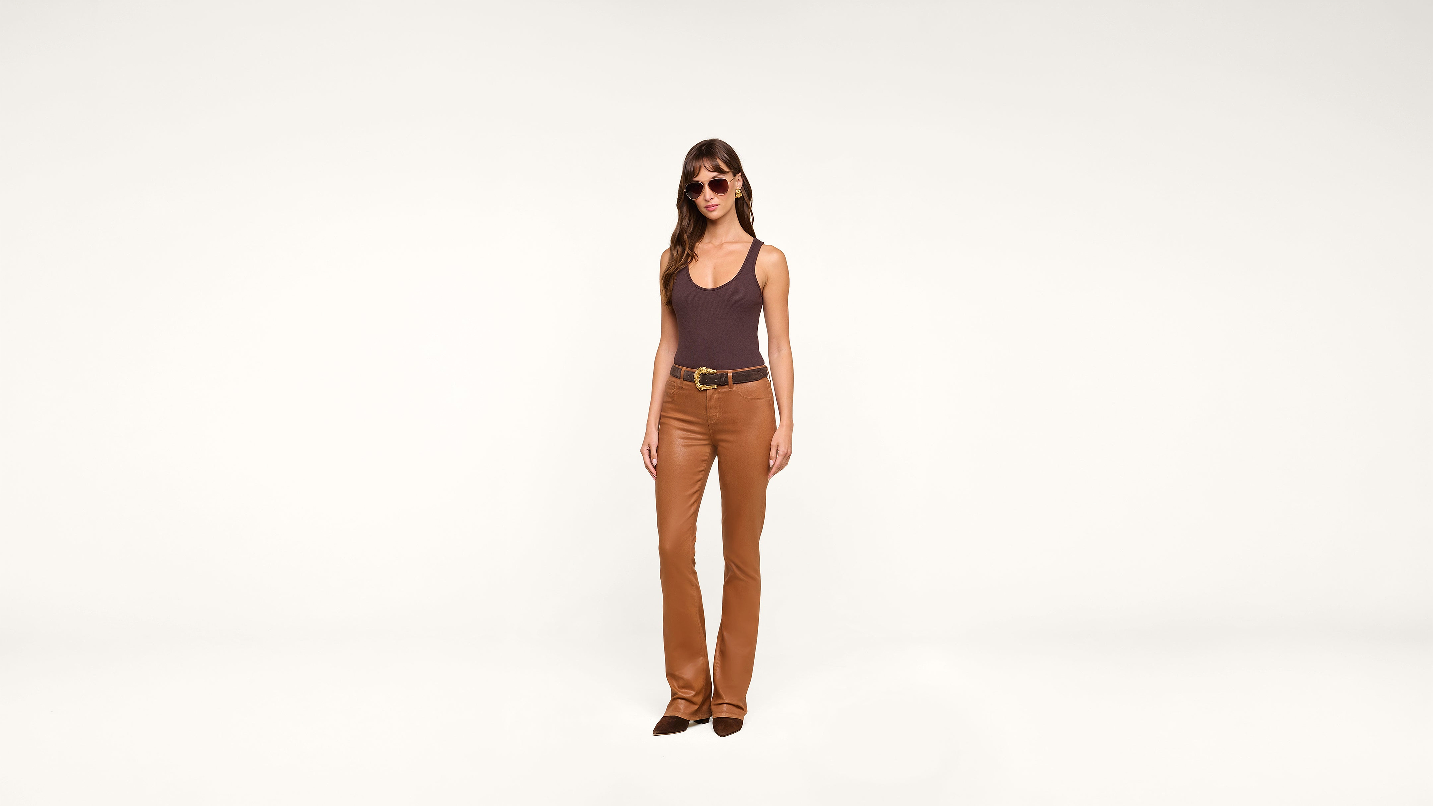 Woman wearing sunglasses, a brown tank top, a black belt, and tan pants stands against a plain white background.
