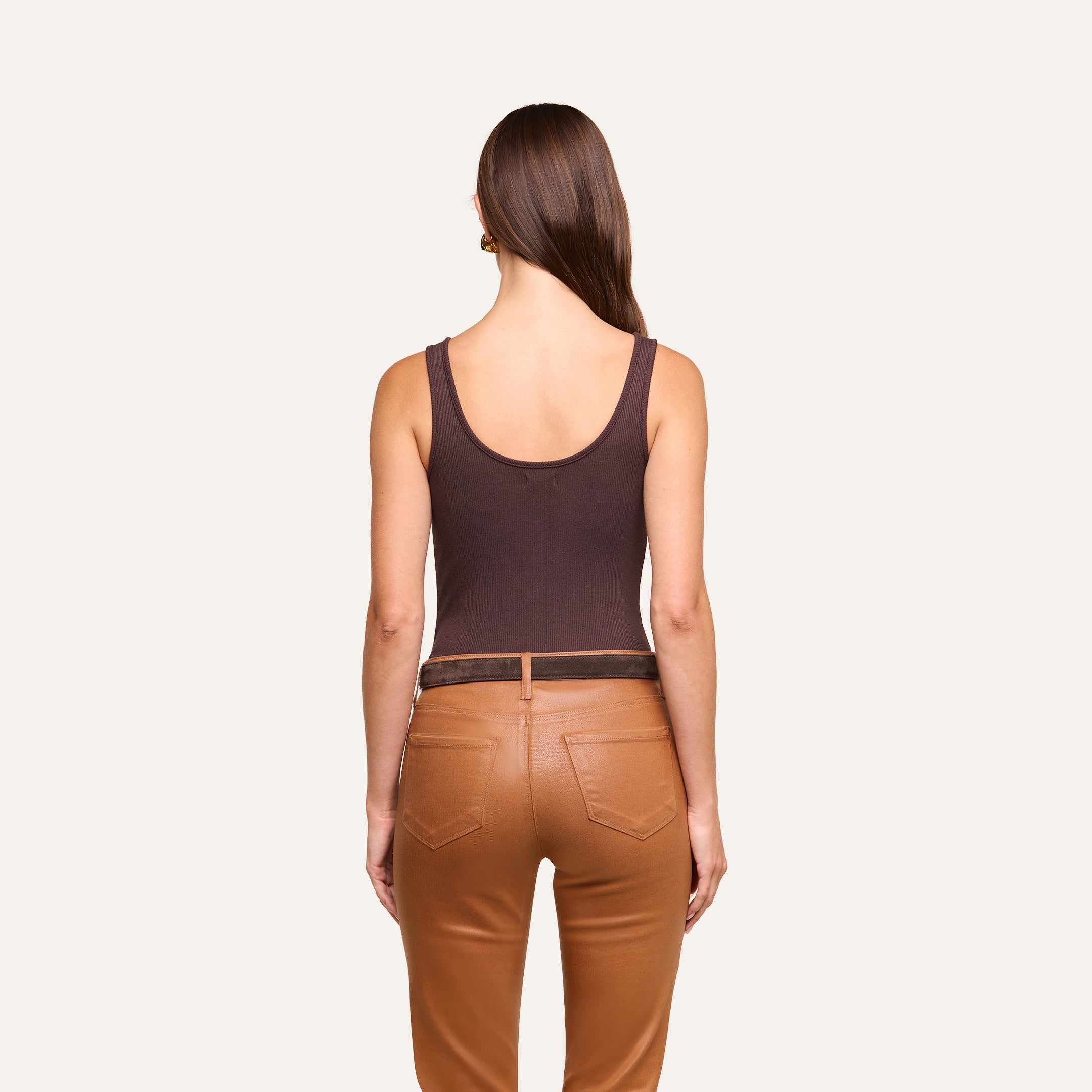A woman stands facing away, wearing a dark brown Nica Scoopneck Bodysuit and tan pants with pockets. Her straight brown hair is down, and she is positioned against a plain light background.