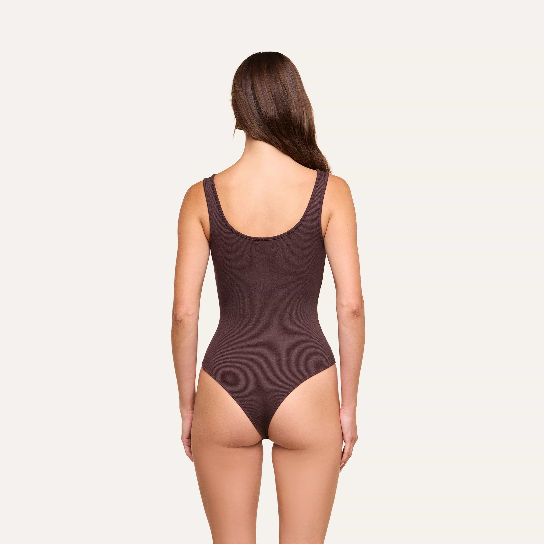 A woman with long brown hair stands facing away, wearing the Nica Scoopneck Bodysuit in dark brown stretch rib fabric against a plain light background.
