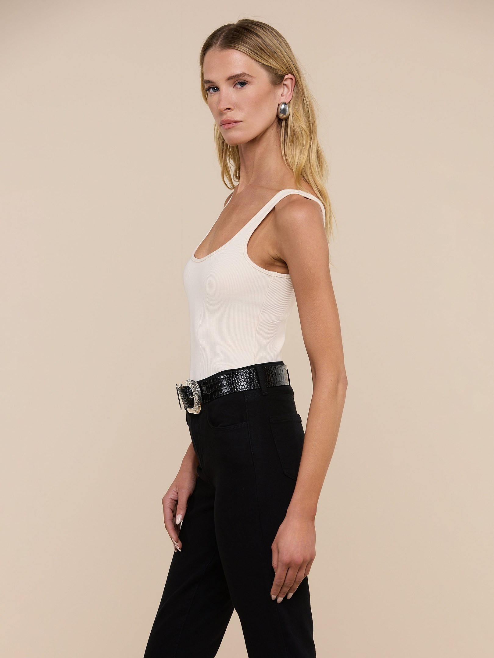 A woman with long blonde hair is wearing the Nica Scoopneck Bodysuit, styled with black pants, a black belt featuring a large buckle, and earrings. She stands against a plain light background, facing left in a relaxed pose.